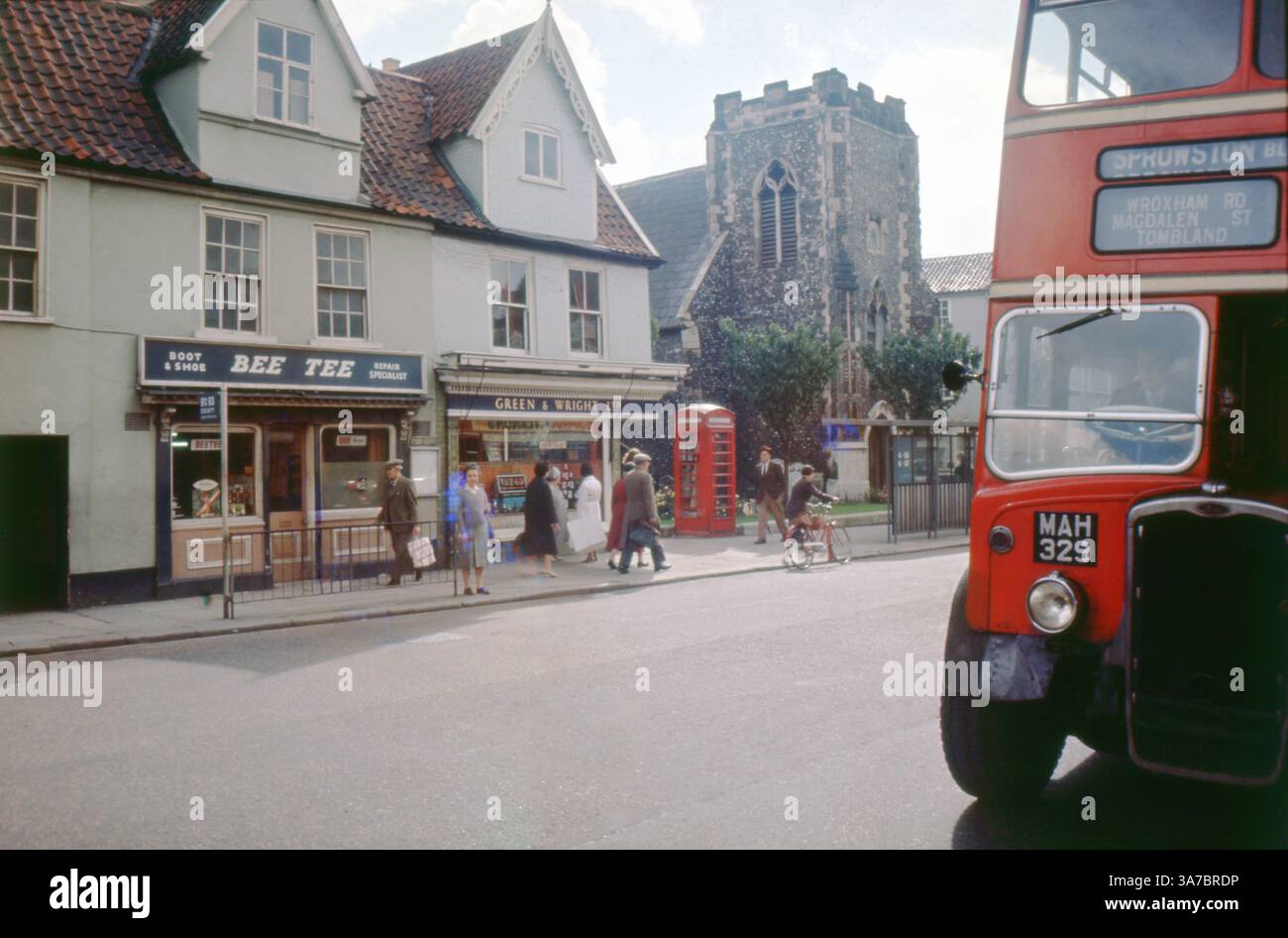Una vivace scena di strada degli anni '1960 in Magdalen Street, Norwich, Norfolk. La gente del posto passeggia davanti a negozi familiari, tra cui BEE TEE Boot & Shoe Repair Specialist e GREEN & WRIGHT LTD Un autobus rosso a due piani contrassegnato con "Sprowston" si prepara a partire, mentre la Chiesa di San Salvatore si erge orgogliosamente sullo sfondo. Foto Stock