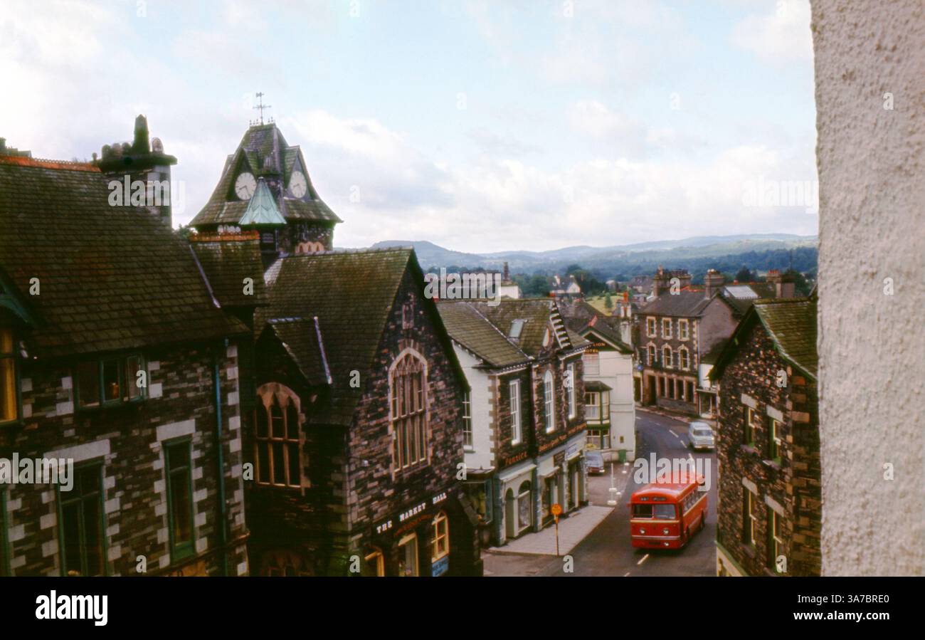 Una pittoresca scena di strada degli anni '1960 di Ambleside, Cumbria, catturata su una pellicola slide da 35 mm. L'iconico Market Hall con la sua impressionante torre dell'orologio si erge orgogliosamente sopra gli edifici con tetto in ardesia della High Street. Un classico autobus rosso attraversa il pittoresco villaggio, incorniciato dalle dolci colline del Lake District in lontananza, offrendo un nostalgico scorcio della vita rurale britannica. Foto Stock