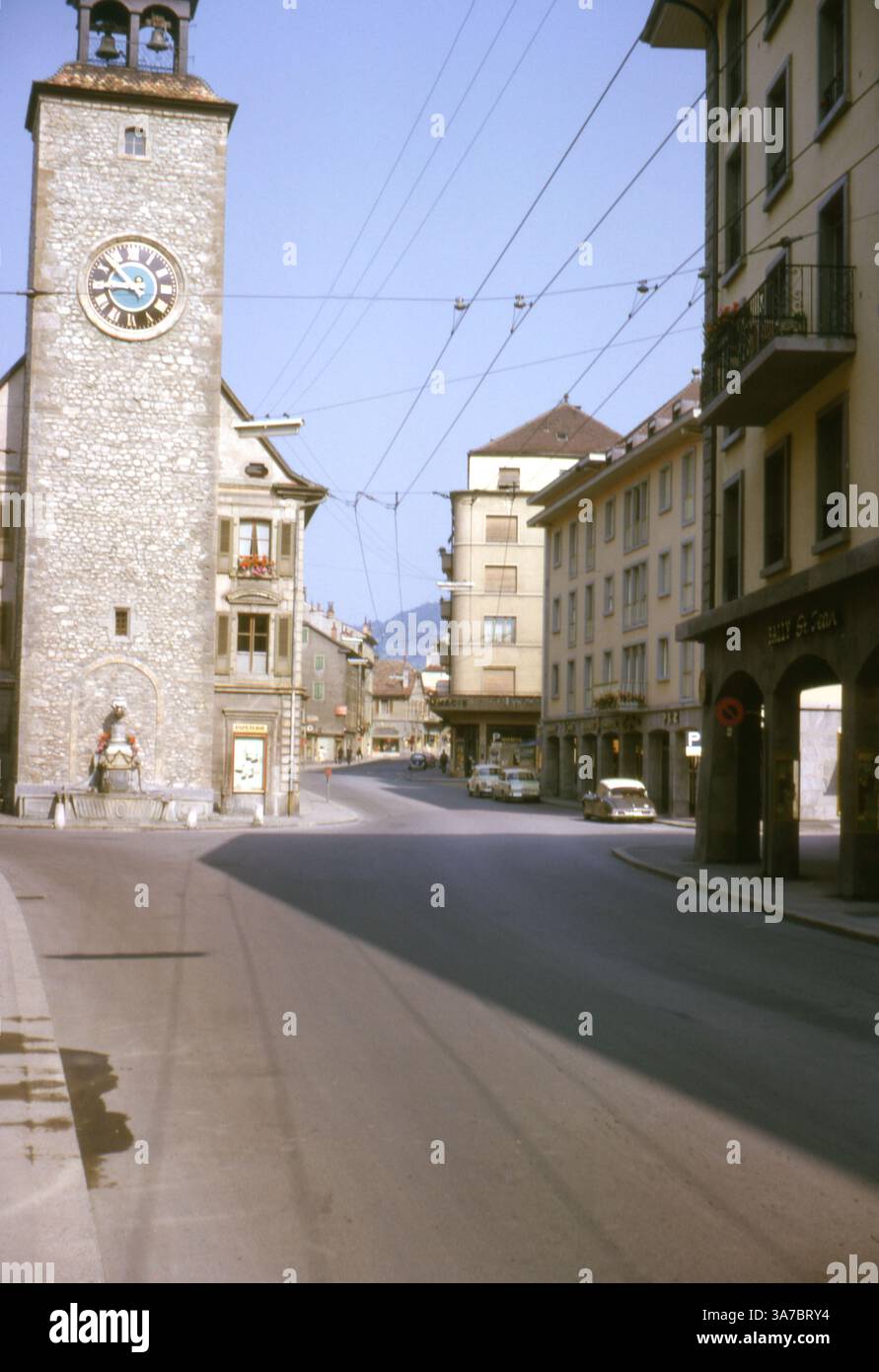 Una tranquilla scena di strada a Vevey, in Svizzera, negli anni '1960, ripresa su pellicola a colori da 35 mm. A dominare la scena c'è l'iconica torre dell'orologio della chiesa di Saint-Martin, la sua facciata in pietra e la faccia blu dell'orologio che si innalza sopra l'architettura della città vecchia. Foto Stock