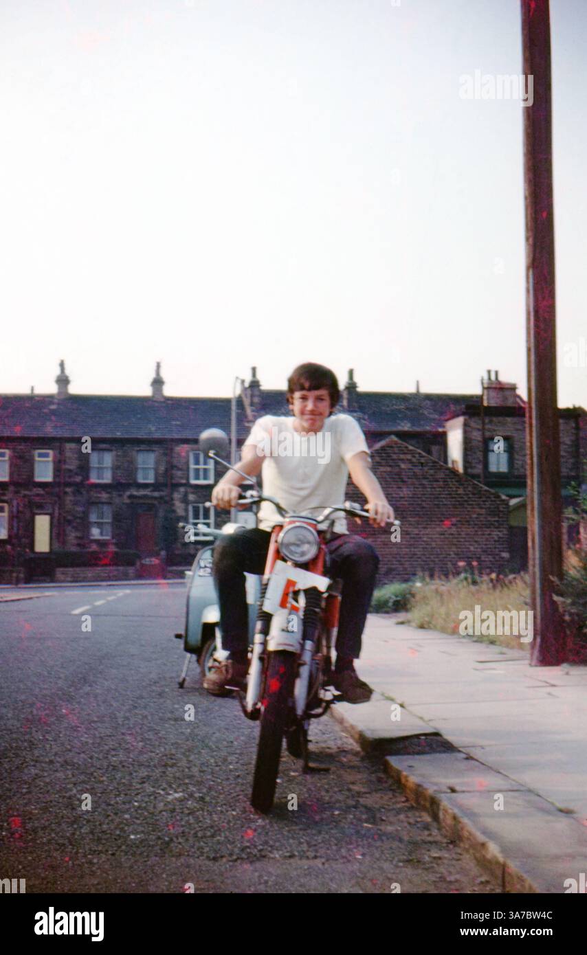 Un giovane uomo siede con fiducia su una motocicletta rossa BSA Bantam in una tranquilla strada inglese del nord durante gli anni '1960 Con un piatto per allievi visibile sulla parte anteriore della moto, la foto cattura un momento nostalgico dei giovani britannici e delle prime esperienze di guida. Foto Stock