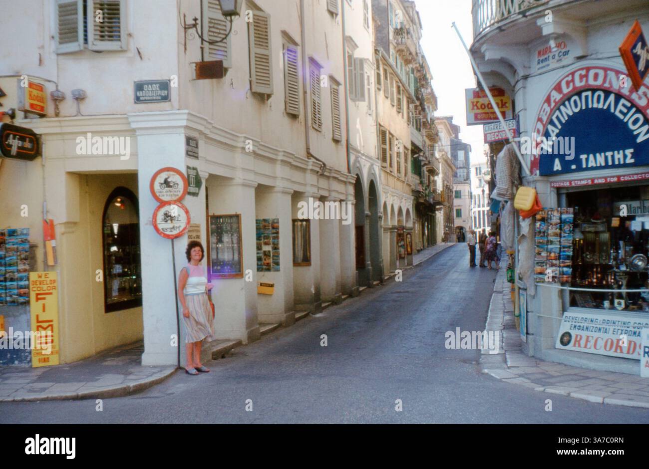 Una vivace scena di strada nella città di Corfù, in Grecia, catturata negli anni '1970 o '1980 su pellicola slide da 35mm. L'immagine mostra i classici edifici con persiane in stile veneziano, banchi da cartolina, insegne Kodak e una donna locale in piedi vicino a un negozio di tabacchi. La strada è costeggiata da piccole aziende e conduce nel cuore della città vecchia Foto Stock