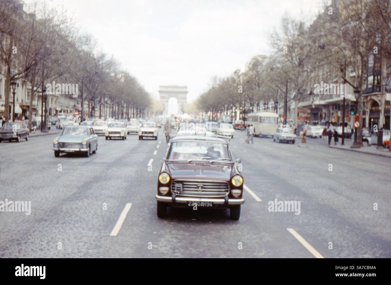 Una classica scena degli anni '1960 lungo gli Champs-Élysées di Parigi, in Francia, caratterizzata da un traffico intenso diretto verso l'Arco di Trionfo. Un taxi Peugeot di colore scuro conduce il flusso di veicoli d'epoca lungo l'ampio viale alberato. Foto Stock