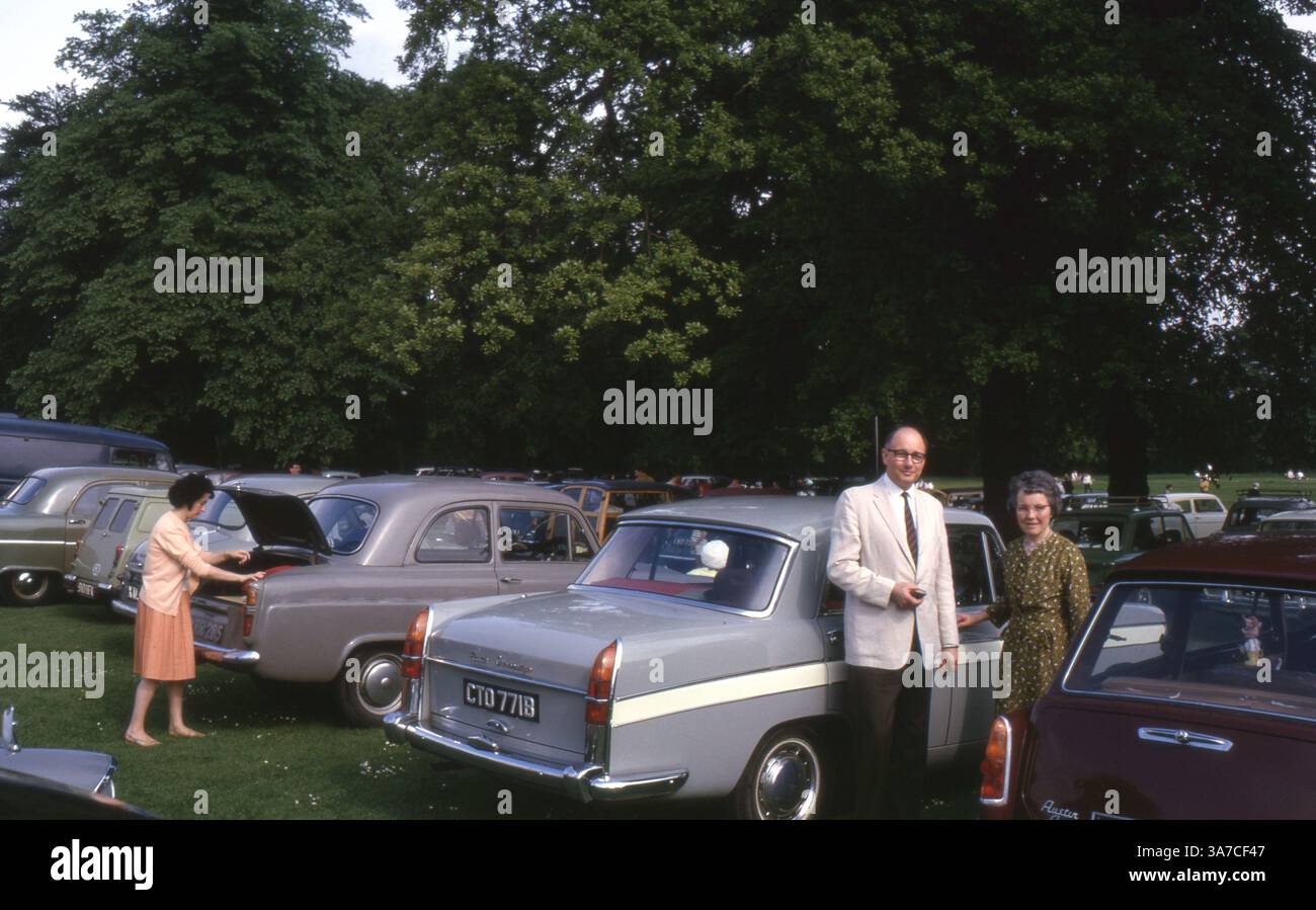 Un uomo e una donna posano per una foto accanto a un Austin Cambridge grigio in un parcheggio in Inghilterra, giugno 1965. La scena cattura una tipica gita estiva della metà degli anni '1960, con le famiglie che scaricano le auto e si preparano per la giornata. Foto Stock