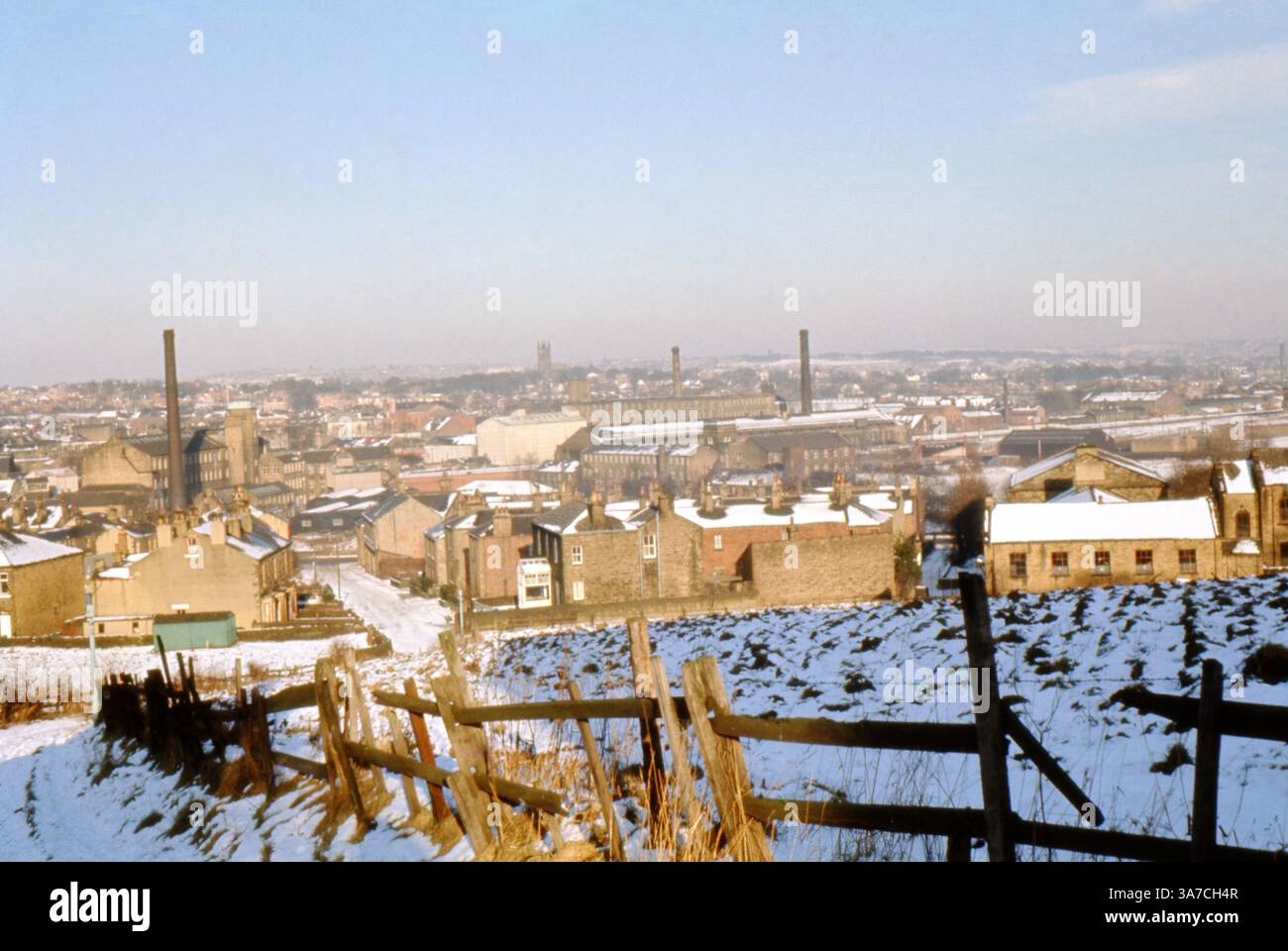 Un panorama invernale di Dewsbury, West Yorkshire, scattato nell'aprile 1969. La neve copre tetti, campi e strade strette in questa storica città industriale. Prominenti camini di mulini si innalzano sopra file di case a schiera e edifici di fabbrica, riflettendo il patrimonio tessile di Dewsbury. La città si estende in lontananza, con la Chiesa di Santa Maria a Mirfield ben visibile sullo skyline. Un'istantanea del paesaggio urbano post-bellico dell'Inghilterra settentrionale in transizione. Foto Stock