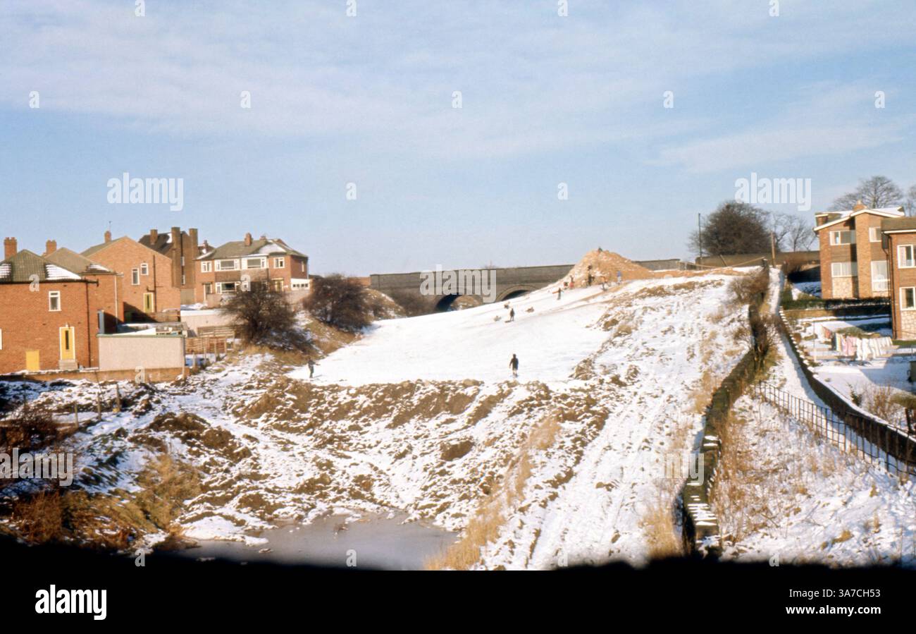 Una scena innevata a Dewsbury, West Yorkshire, catturata nell'aprile 1969. I bambini sono visti scendere in slitta lungo una collina circondata da abitazioni del dopoguerra, con un viadotto ferroviario in pietra in lontananza. L'immagine riflette un momento di svago durante una nevicata insolitamente tardiva, offrendo uno sguardo sulla vita in una città industriale del nord durante gli anni '1960 Foto Stock