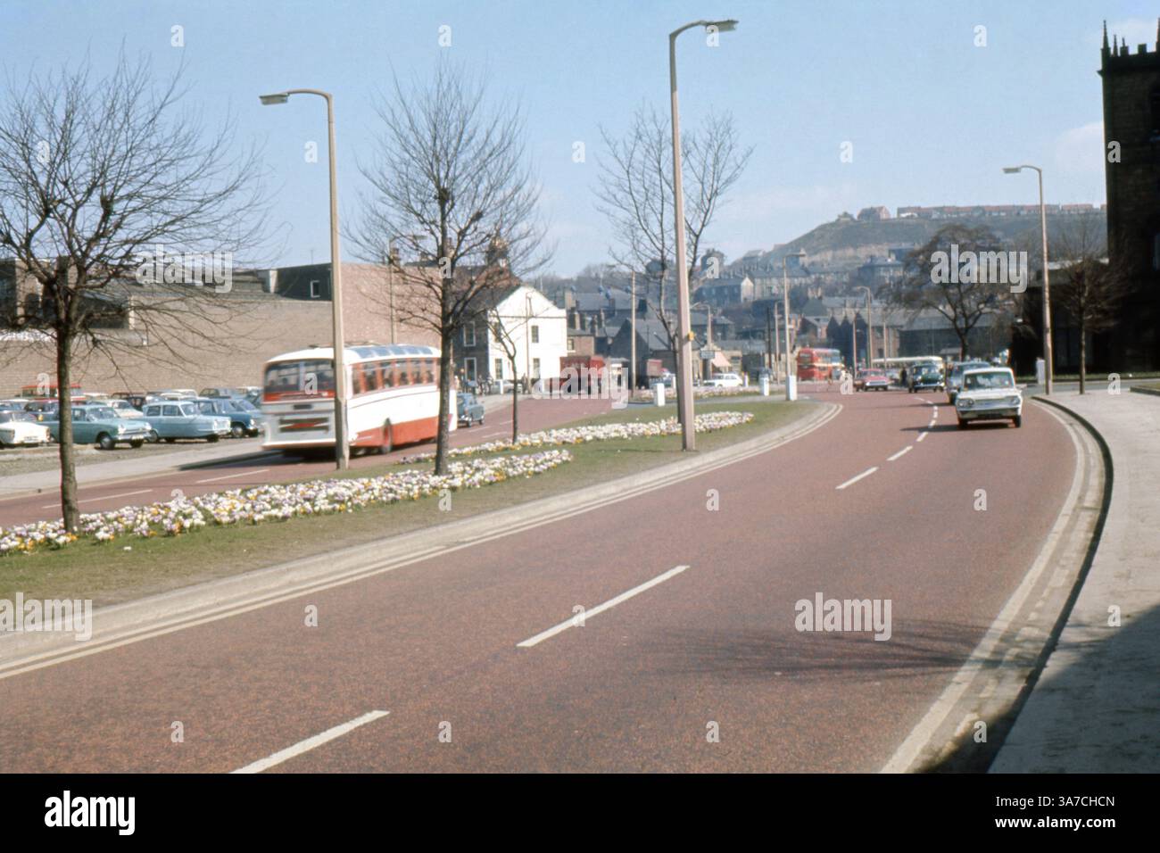 La fotografia originale del 1969 cattura Huddersfield Road a Dewsbury, nel West Yorkshire, in un giorno di primavera di sole. La strada è fiancheggiata da veicoli classici e dai confini fioriti, con autobus, automobili e negozi locali visibili in lontananza. L'immagine offre uno sguardo nostalgico sulla vita cittadina britannica della fine degli anni '1960, mostrando una fiorente scena urbana con colline ed edifici residenziali sullo sfondo. Foto Stock