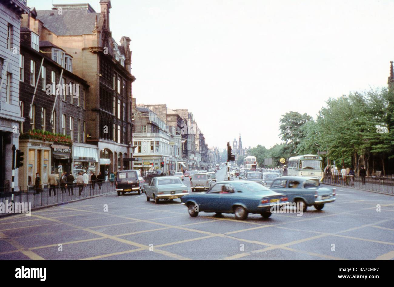 Una vivace scena di strada di Edimburgo degli anni '1970 cattura la vivacità di Princes Street, una delle strade più iconiche della città. Veicoli d'epoca, autobus a due piani e pedoni riempiono l'ambiente urbano, con la sagoma del monumento Scott che si innalza in lontananza. Traffico proveniente dalla destra su Lothian Road. Foto Stock