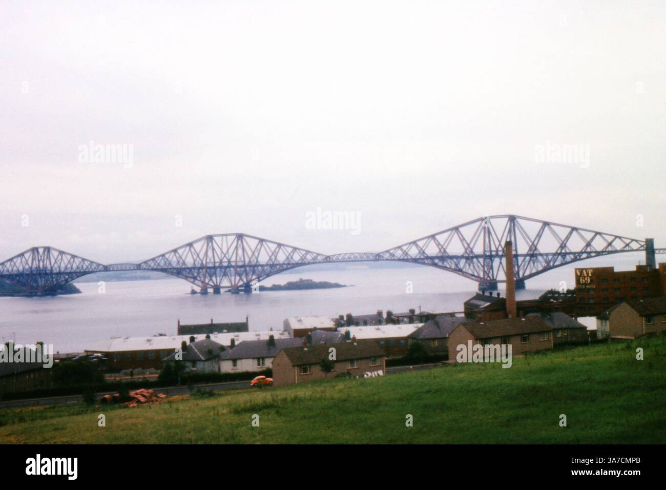 Una vista del 1970 dell'iconico Forth Bridge che attraversa il Firth of Forth in Scozia, con un primo piano di tetti residenziali e un edificio industriale che pubblicizza il whisky VAT 69. Foto Stock