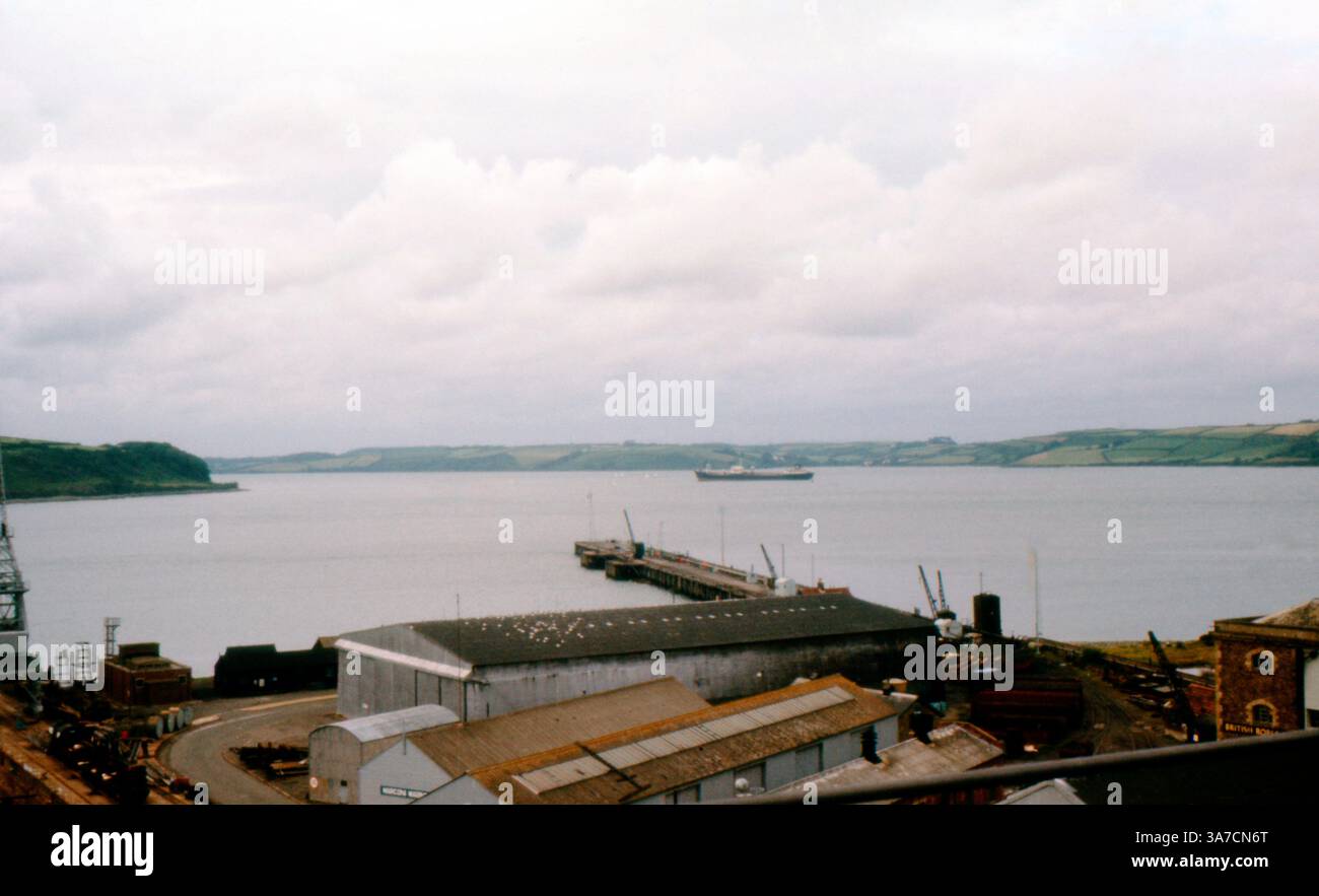 Una vista sui moli di Falmouth in Cornovaglia durante gli anni '1970, mostrando gli edifici del cantiere navale, le infrastrutture portuali e una nave ancorata nell'estuario. La scena mette in evidenza il patrimonio industriale marittimo della città e la sua posizione strategica sull'estuario del Fal, con campi ondulati visibili lungo l'acqua. Foto Stock