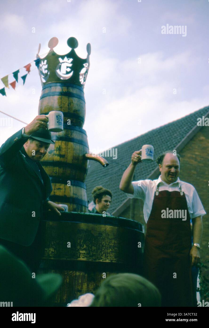 Due uomini alzano le canne con un brindisi allegro accanto a un torreggiante barile di birra a corona al 1974 Schützenfest di Aligse, Germania. Uno indossa l'abbigliamento tradizionale del birraio e l'altro un abito e un cappello, che incarnano lo spirito comune e l'orgoglio culturale di questo festoso raduno rurale. Foto Stock