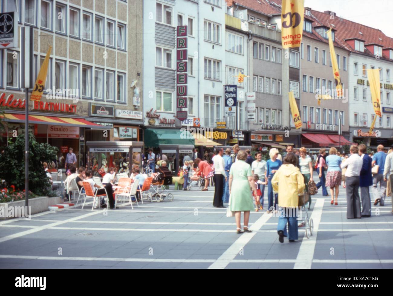 Una vivace zona pedonale a Hildesheim, Germania, nell'agosto 1976. Gli amanti dello shopping e le famiglie apprezzano l'atmosfera vivace, con posti a sedere all'aperto, vetrine e cartelli che riflettono la vita cittadina tedesca degli anni '1970. Le aziende visibili includono un parrucchiere (“Friseur”), caffetterie, uno stand di bratwurst e una filiale di Eduscho, un popolare rivenditore di caffè. Foto Stock