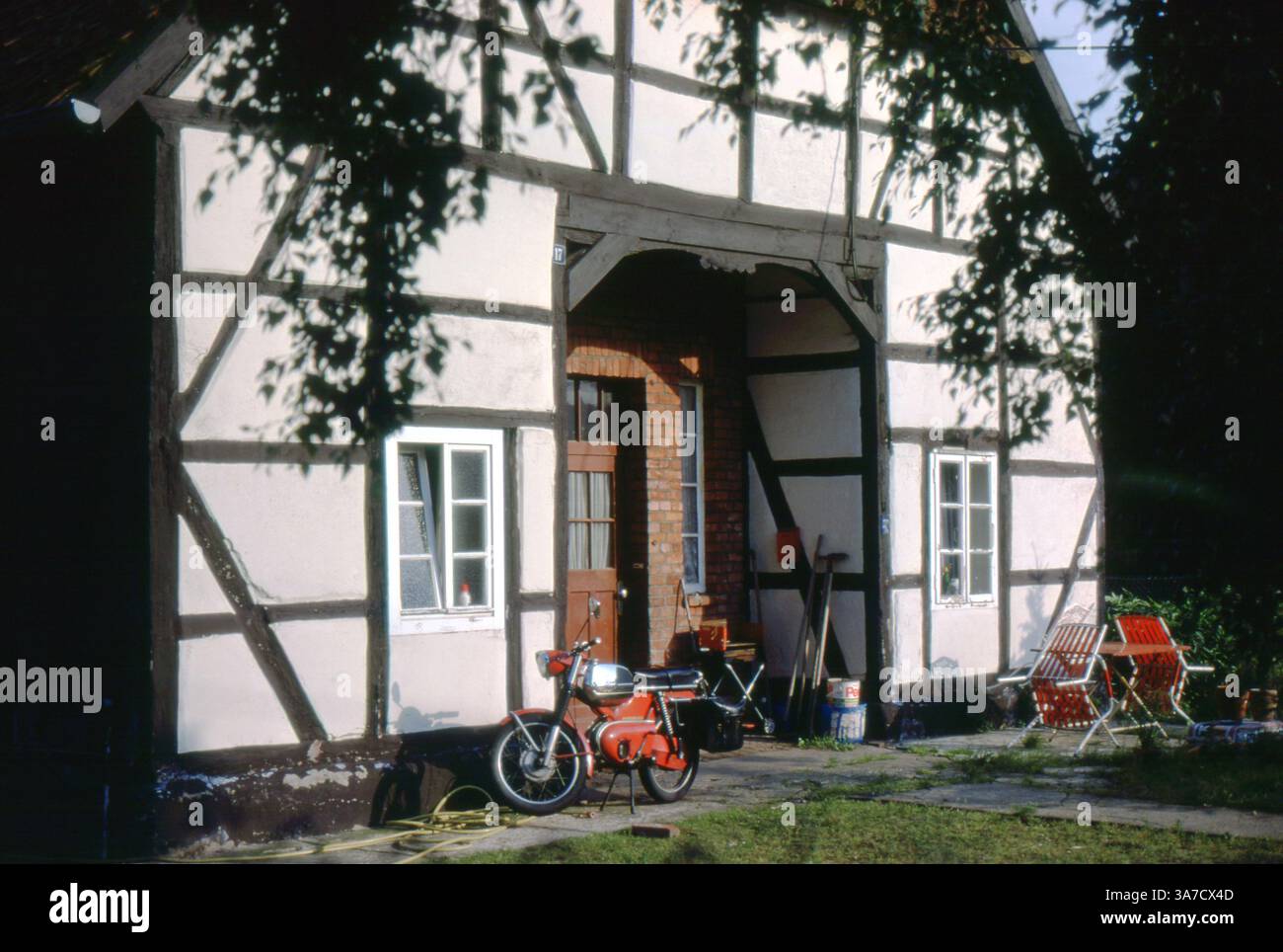 Una fattoria tradizionale a Sievershausen, in Germania, catturata nel luglio 1979. Una moto rossa d'epoca è parcheggiata all'ingresso, mentre le sedie rosse e gli attrezzi da giardino suggeriscono un'atmosfera rurale vissuta. Foto Stock