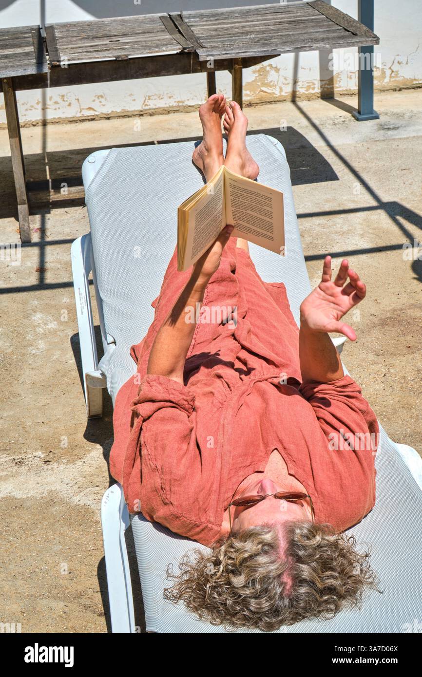Donna anziana che si gode una giornata rilassante leggendo un libro mentre si sdraia su una sedia a sdraio al sole Foto Stock