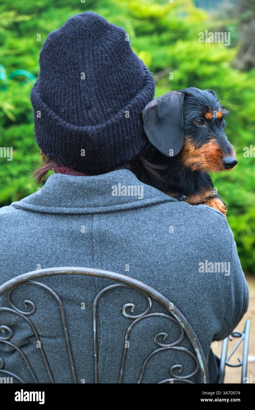 Donna con berretto seduta su una sedia di metallo, che tiene il suo cane con i capelli in un giardino verde Foto Stock