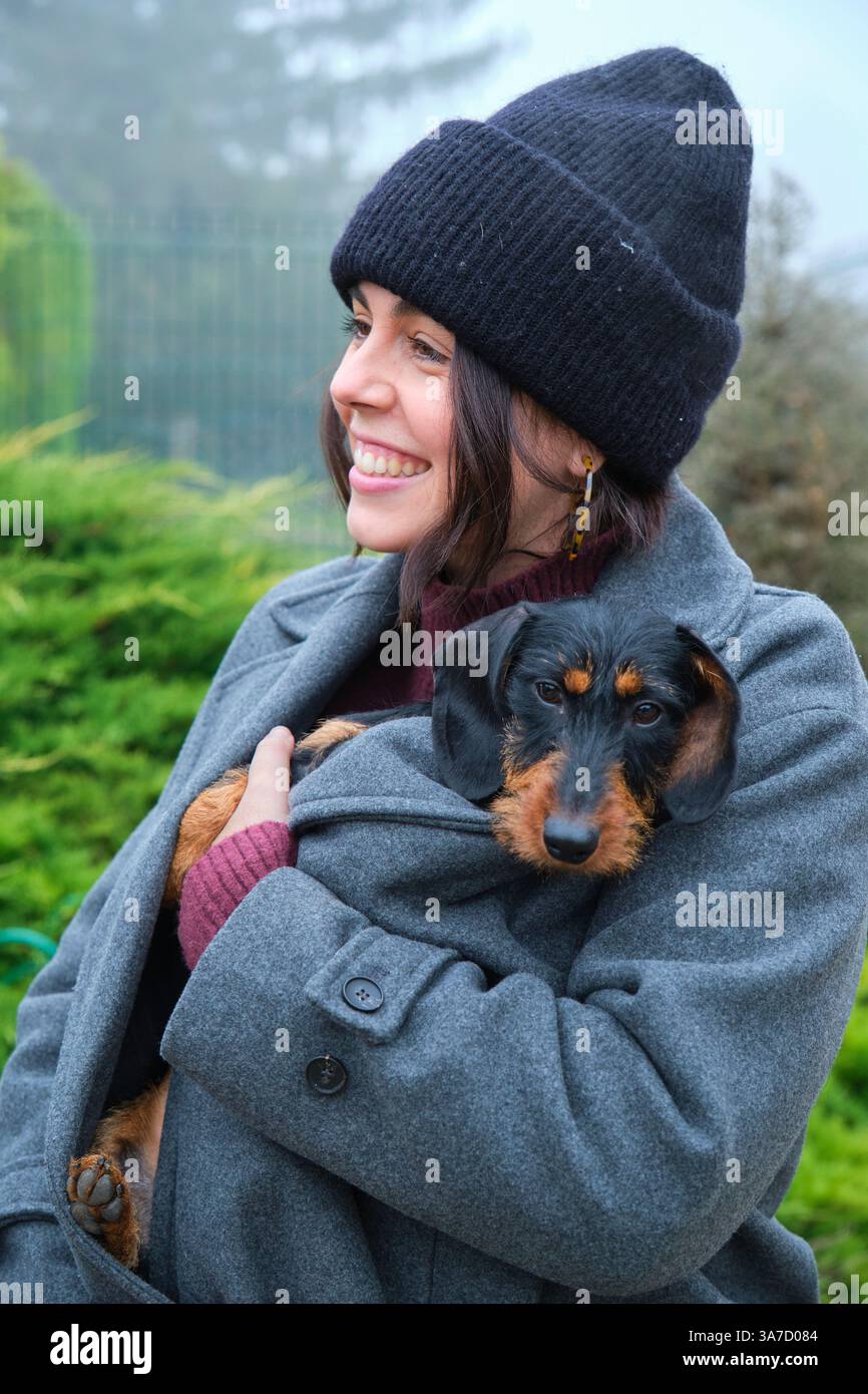 La giovane donna felice con un berretto nero tiene il suo cane all'interno del suo caldo cappotto grigio durante una fredda giornata nebbiosa Foto Stock