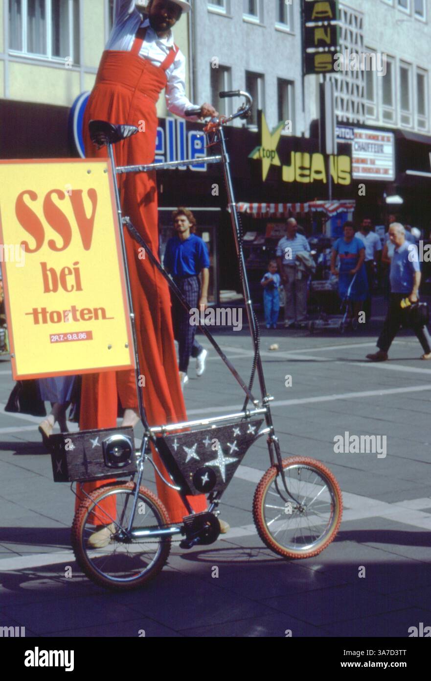 Un vivace artista di strada su palafitte pubblicizza una vendita stagionale presso i grandi magazzini Horten di Hildesheim, Germania, durante l'estate del 1986. Vestito con tute di rosso brillante, l'intrattenitore cavalca una bicicletta alta comicamente modificata lungo una trafficata strada pedonale per lo shopping, catturando l'attenzione con un cartello giocoso e un'esibizione stravagante Foto Stock