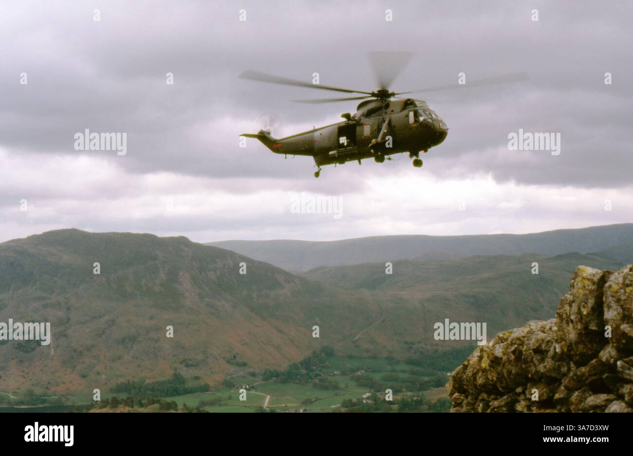 Un elicottero Westland Sea King dell'esercito britannico si libra in alto sopra il Lake District durante un'esercitazione militare o un addestramento di soccorso in montagna nel maggio 1987. Catturata a mezz'aria con le conchiglie e le valli della Cumbria che si estendono sotto, questa immagine riflette il terreno impegnativo e le sfide operative affrontate dagli equipaggi militari in ambienti montuosi. Foto Stock