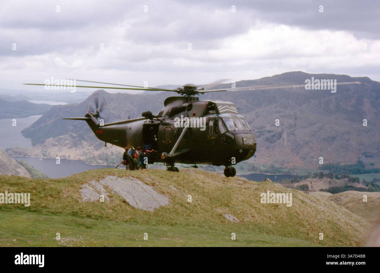 Un elicottero Westland Sea King dell'esercito britannico conduce un'operazione di addestramento o salvataggio in montagna nel Lake District, maggio 1987. L'aereo è raffigurato a metà volo come personale con pacchi e equipaggiamento a bordo su terreni accidentati che si affacciano su un lago. Foto Stock