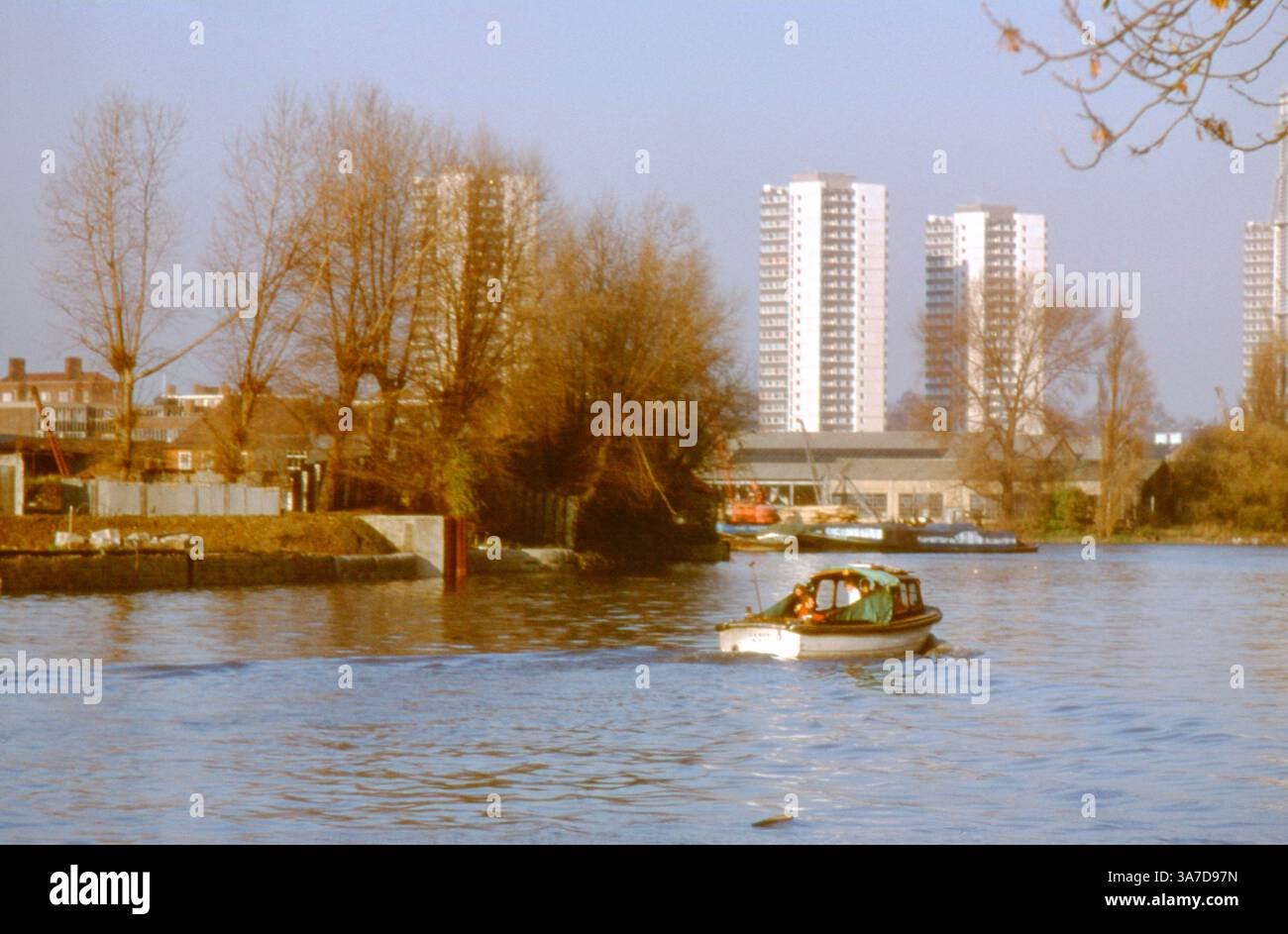 Una piccola barca naviga oltre il fiume Brentford sul Tamigi, con blocchi a torre degli anni '1990 e l'industria fluviale sullo sfondo. Foto Stock
