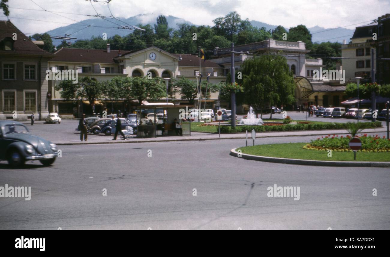Una vista del 1960 della stazione ferroviaria di Vevey nel cantone di Vaud, in Svizzera. Questo vivace paesaggio urbano cattura il piazzale della stazione con auto parcheggiate, un chiosco e una fontana centrale, il tutto sullo sfondo dell'architettura tradizionale e delle colline ricoperte di alberi. Foto Stock
