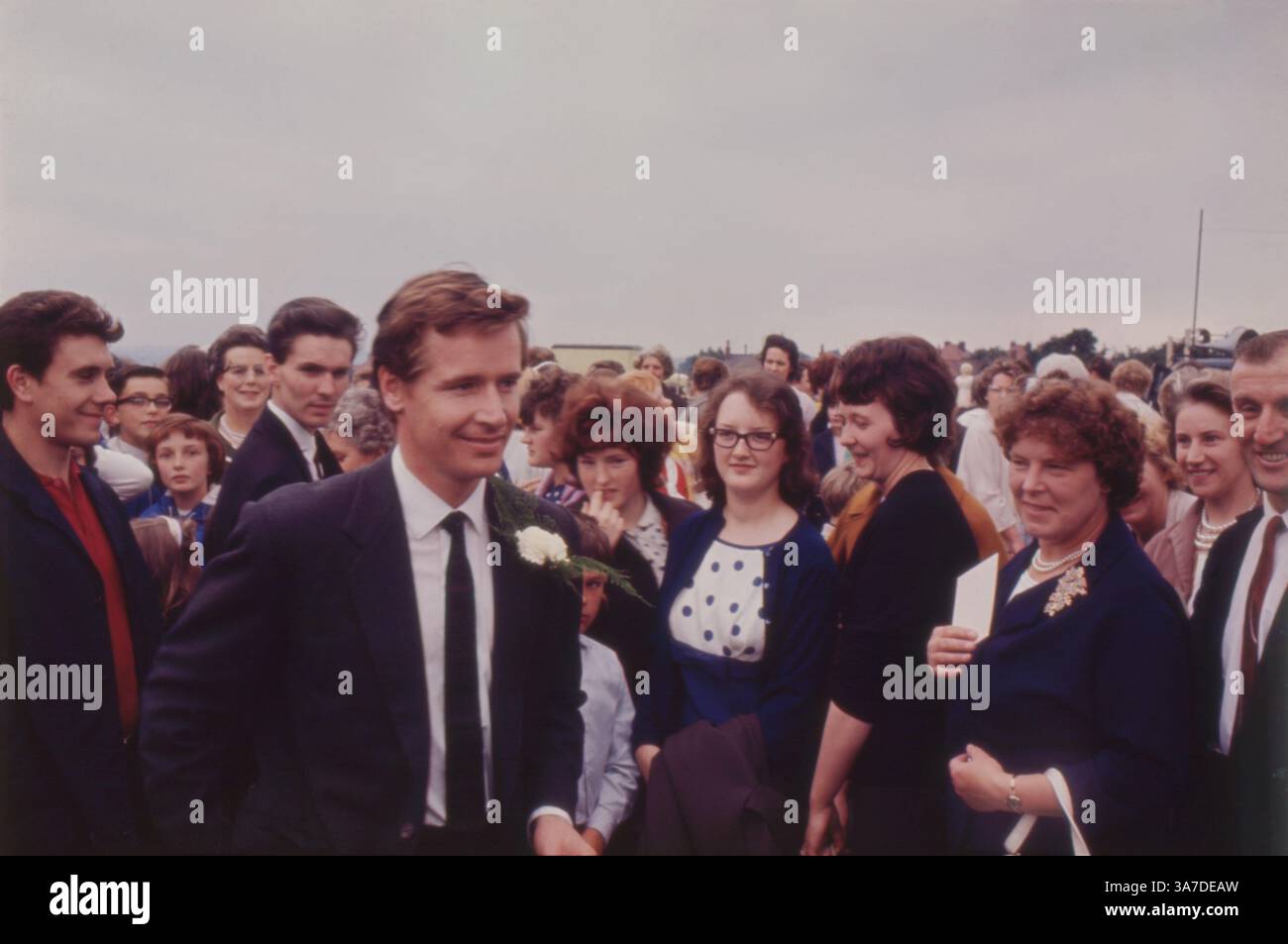 L'attore William (Bill) Roache, meglio conosciuto per il suo ruolo in Coronation Street, è visto partecipare a un evento pubblico all'aperto negli anni '1960 Vestito con una tuta scura con un fiore all'occhiello di garofano bianco, Roache si mescola con una folla di fan, circondato da donne, bambini e spettatori. Foto Stock