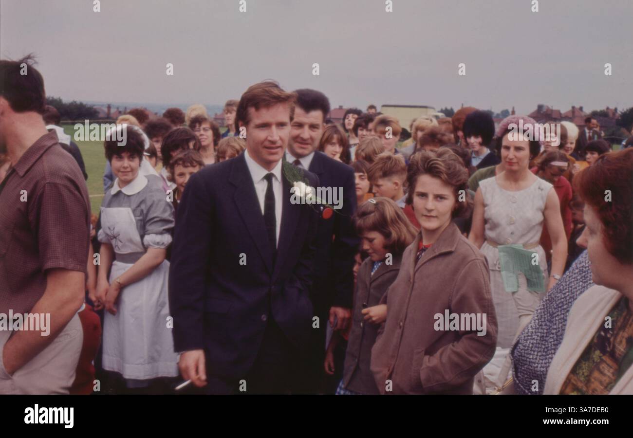 L'attore William (Bill) Roache, meglio conosciuto per il suo ruolo in Coronation Street, è visto partecipare a un evento pubblico all'aperto negli anni '1960 Vestito con una tuta scura con un fiore all'occhiello di garofano bianco, Roache si mescola con una folla di fan, circondato da donne, bambini e spettatori. Foto Stock