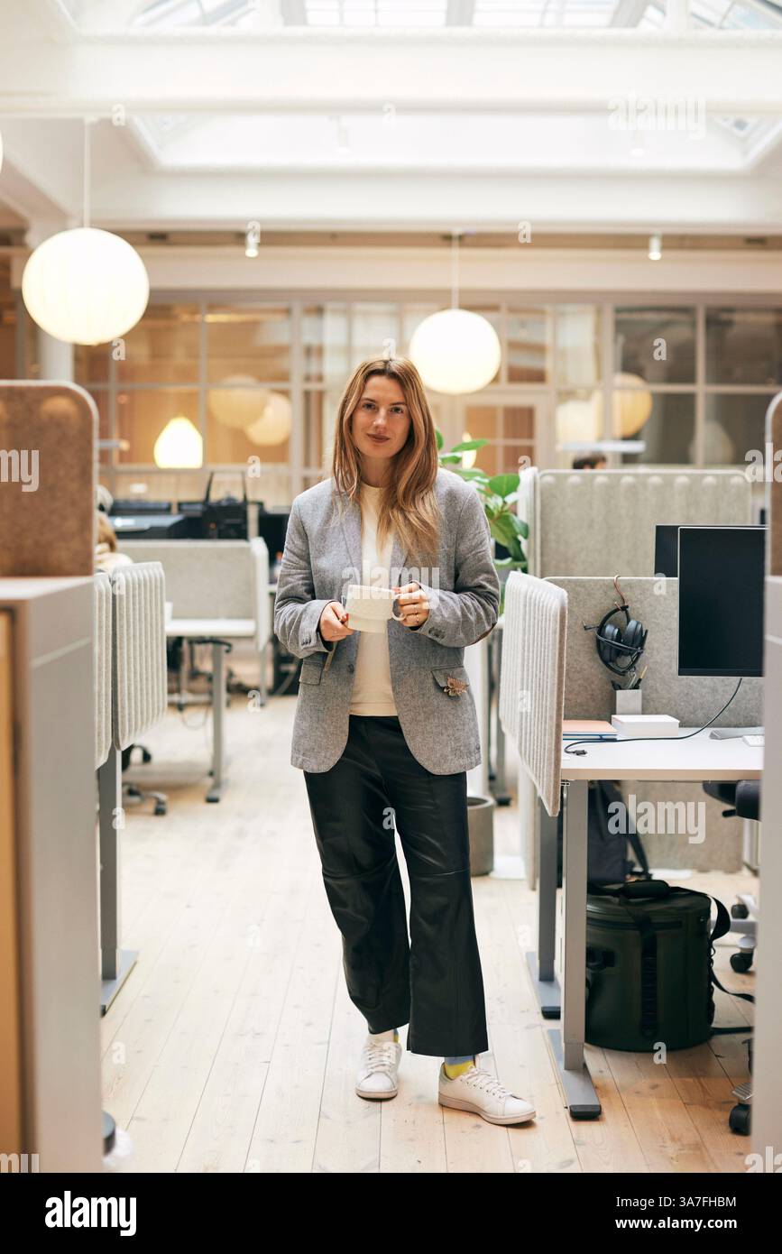 Ritratto a tutta lunghezza di una donna sicura di sé che tiene la tazza mentre si trova in ufficio Foto Stock