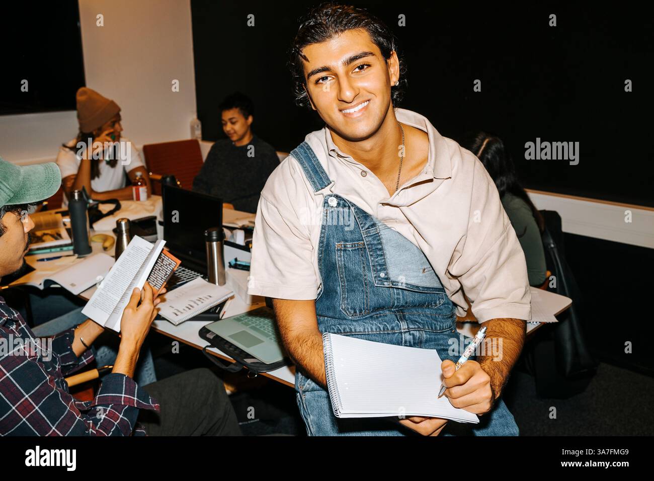 Ritratto di un giovane felice che indossa il pettorale tenendo in mano il diario mentre si siede sul tavolo in classe Foto Stock