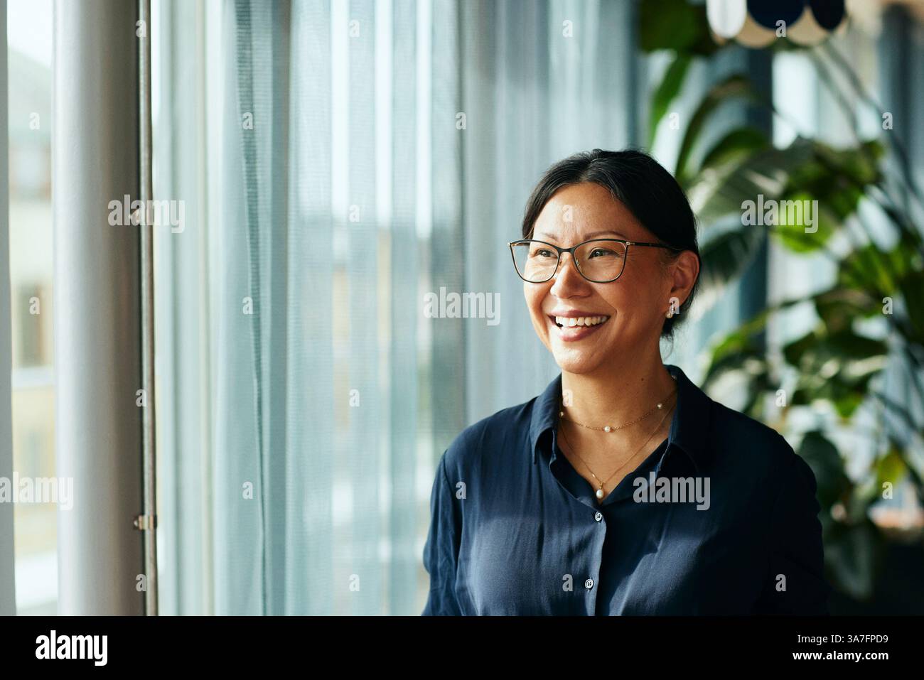 Donna d'affari felice e matura con occhiali che guarda dalla finestra dell'ufficio Foto Stock