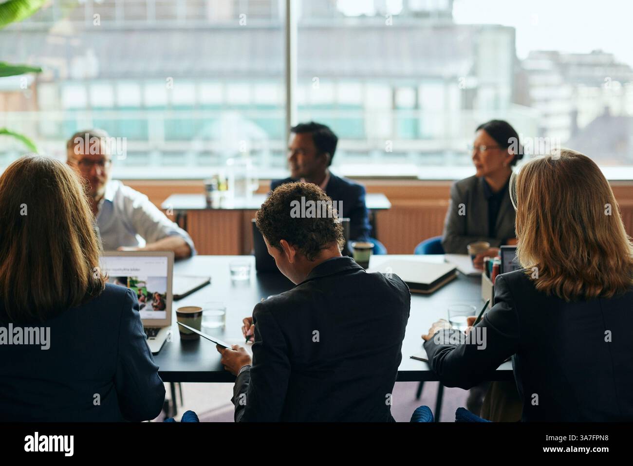 Vista posteriore di una donna d'affari che scrive appunti mentre siede con i colleghi al tavolo da conferenza nella sala riunioni dell'ufficio Foto Stock