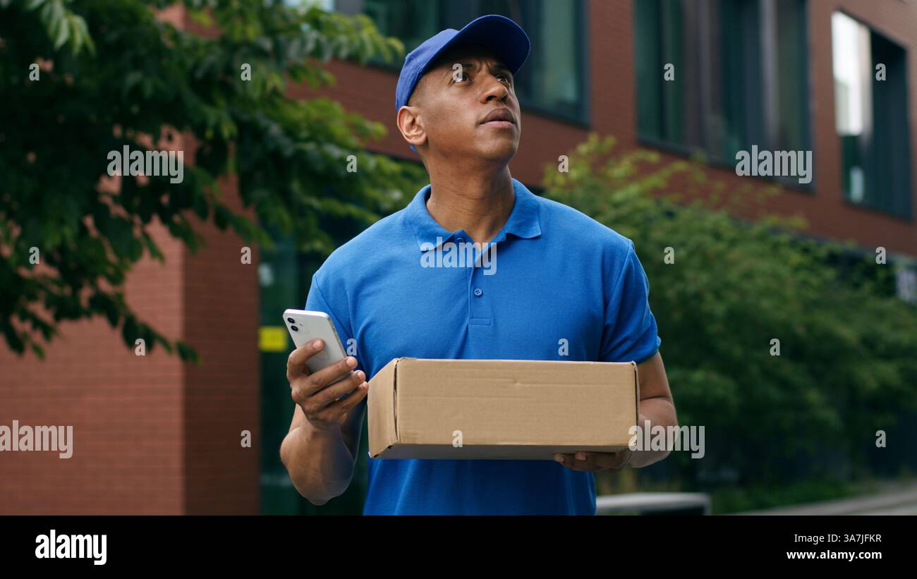 Corriere afroamericano confuso guardare la mappa GPS del telefono cellulare ricerca online indirizzo di posizione guardando intorno tenendo in mano il corriere postale Foto Stock