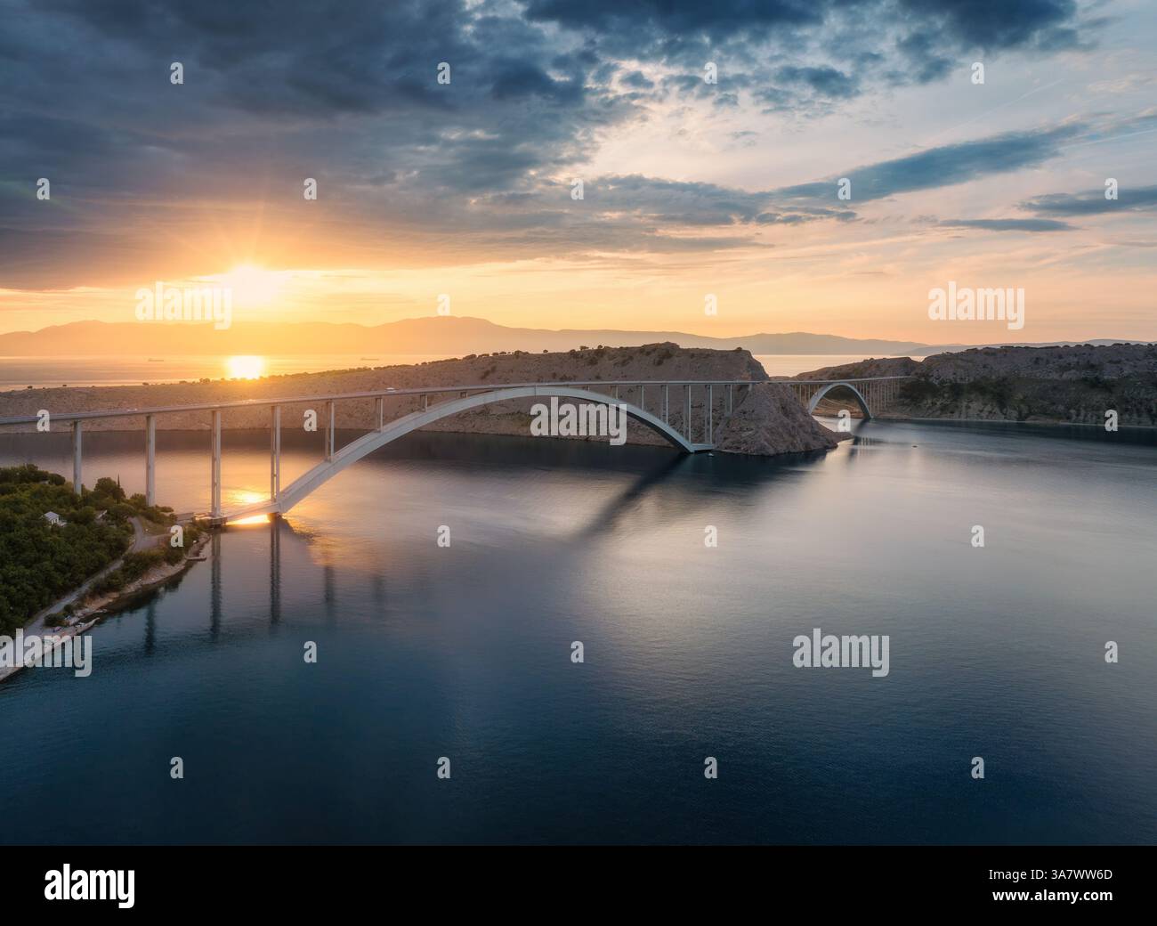 Vista aerea del ponte e del mare blu al tramonto colorato. Moderna isola del ponte di Krk durante l'ora d'oro, montagne, rocce, riflessi nell'acqua e nuvolose Foto Stock