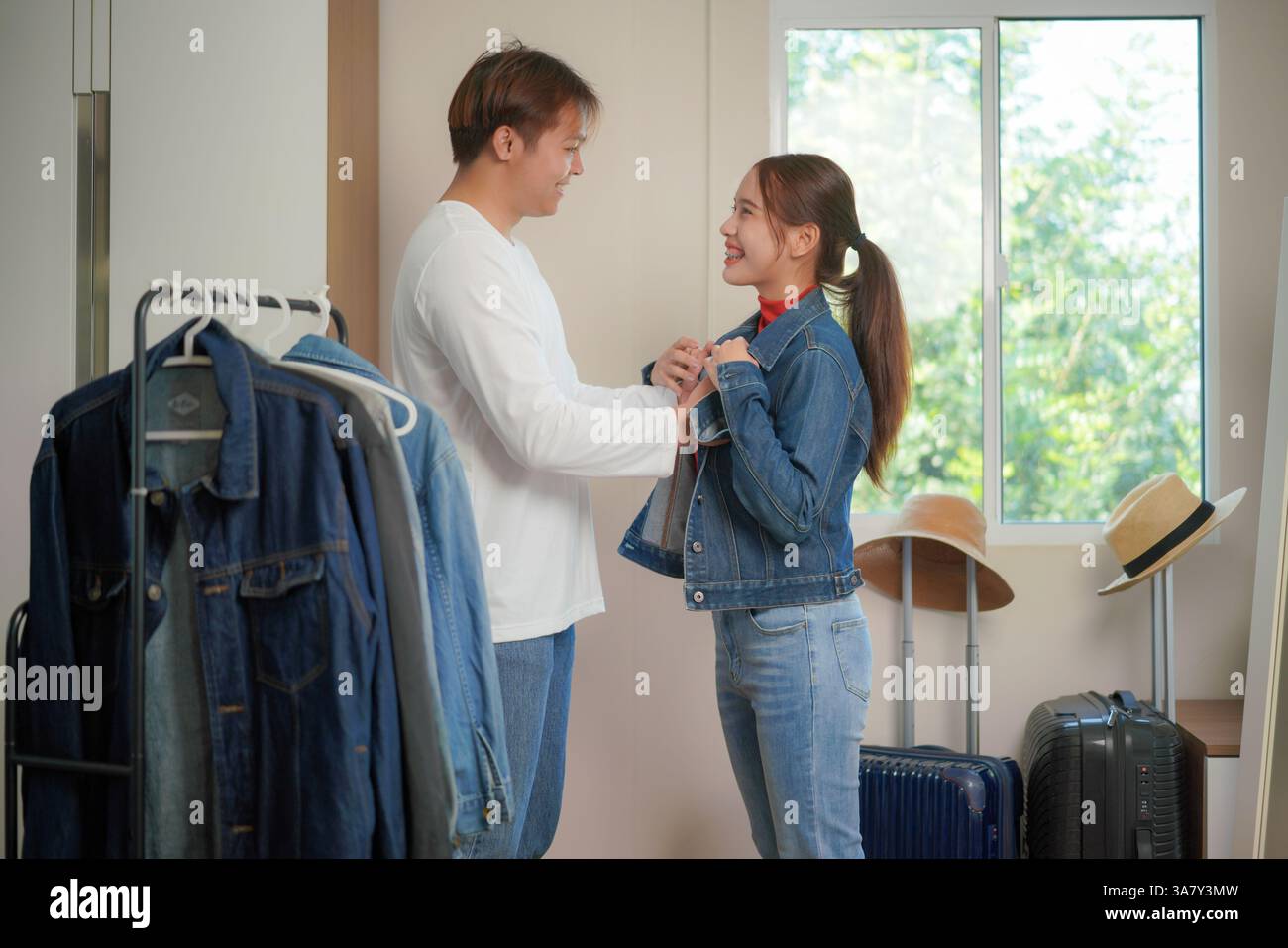 il giovane uomo e la donna stanno in una stanza ben illuminata, sorridendo l'un l'altro mentre finalizzano la sua giacca in denim. Con un portapacchi e valigie nel dorso Foto Stock