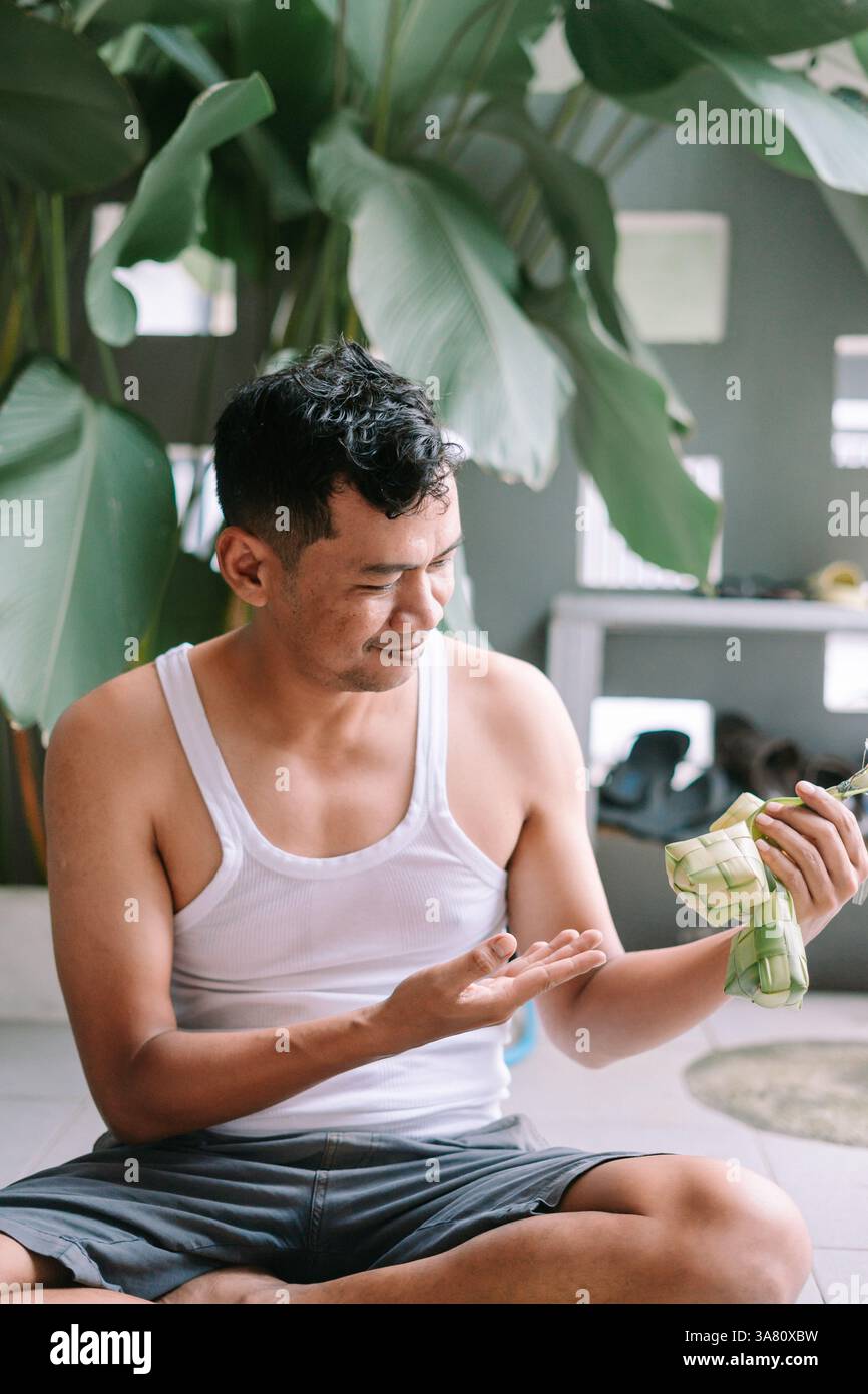 Un uomo che indossa una canotta bianca tiene in mano Ketupat, una tradizionale torta di riso del sud-est asiatico. Questo cibo viene spesso servito durante la celebrazione di Eid al-Fitr Foto Stock