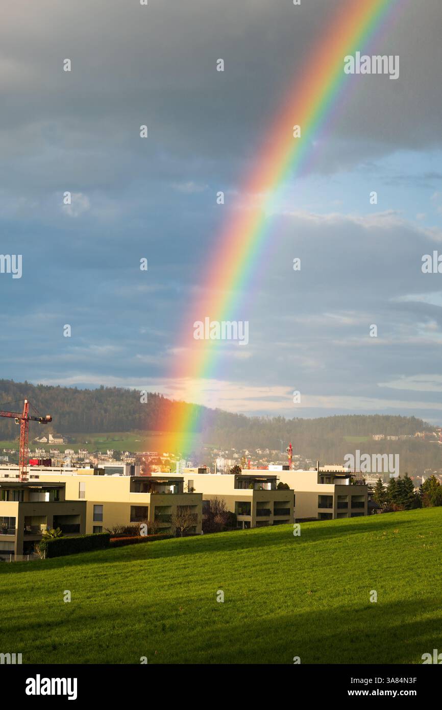 Un bellissimo arcobaleno che si abbatte dietro i piccoli edifici della città dopo la tempesta. Campo verde in primo piano, cielo nuvoloso, nessuna gente. Foto Stock