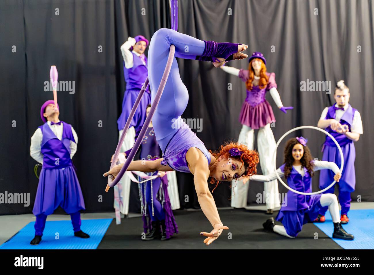 Redhead acrobat si esibisce su un cerchio aereo, circondato da giocolieri e artisti circensi, mettendo in risalto il lavoro di squadra e l'arte Foto Stock Redhead acrobat si esibisce su un cerchio aereo, circondato da giocolieri e artisti circensi, mettendo in risalto il lavoro di squadra e l'arte Foto Stock