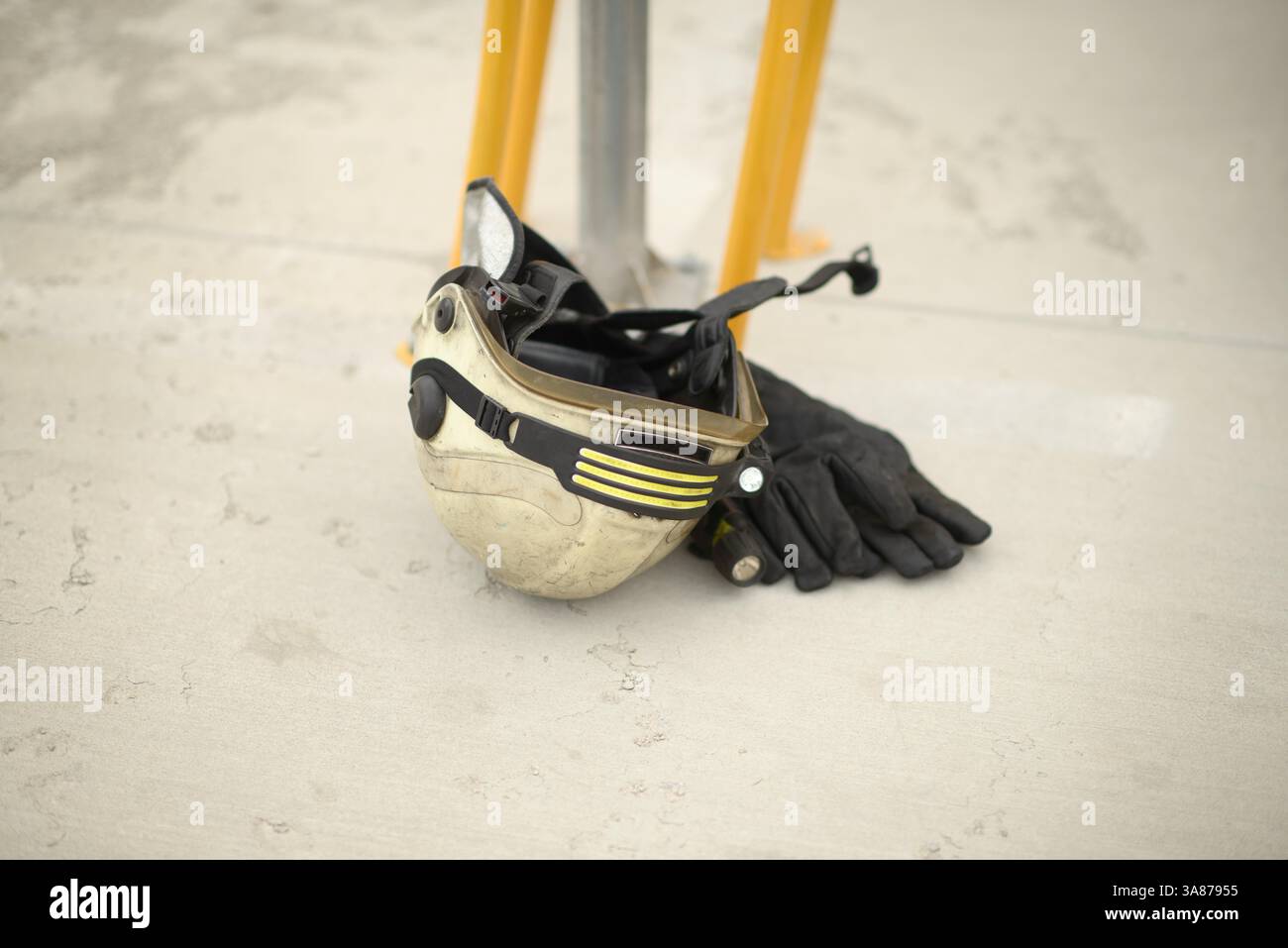 Un casco da vigile del fuoco indossato con strisce riflettenti e guanti protettivi neri si trova su una superficie di cemento accanto a un palo strutturale giallo, pronto per l'uso Foto Stock