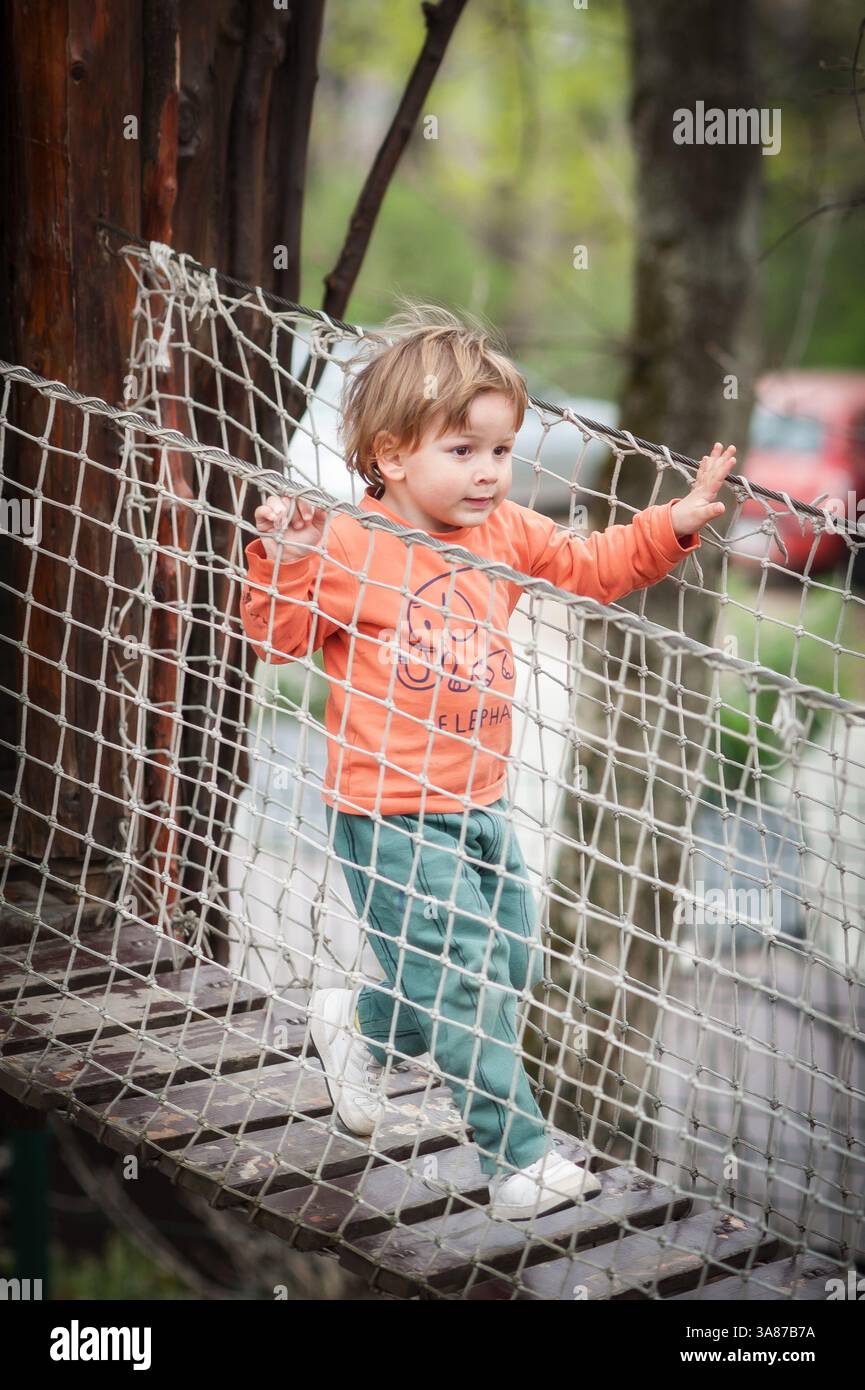 Un bambino cammina con sicurezza attraverso un ponte di corda in un parco giochi, circondato da vegetazione lussureggiante e strutture in legno, godendosi una giornata luminosa all'aperto Foto Stock