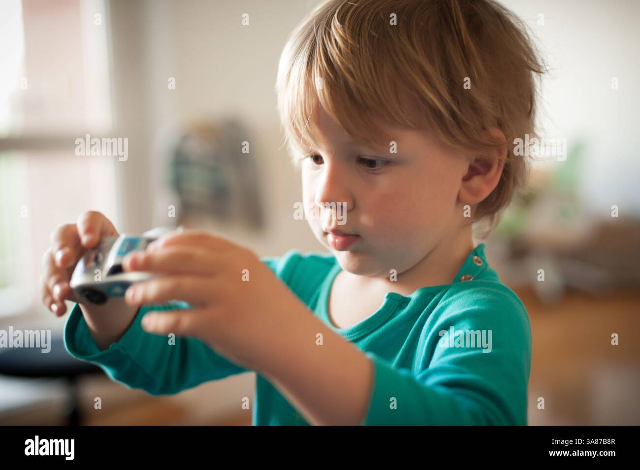 Un bambino piccolo, concentrato e curioso, esplora un'auto giocattolo seduto al chiuso. La luce calda illumina la stanza, creando un'atmosfera allegra. Foto Stock