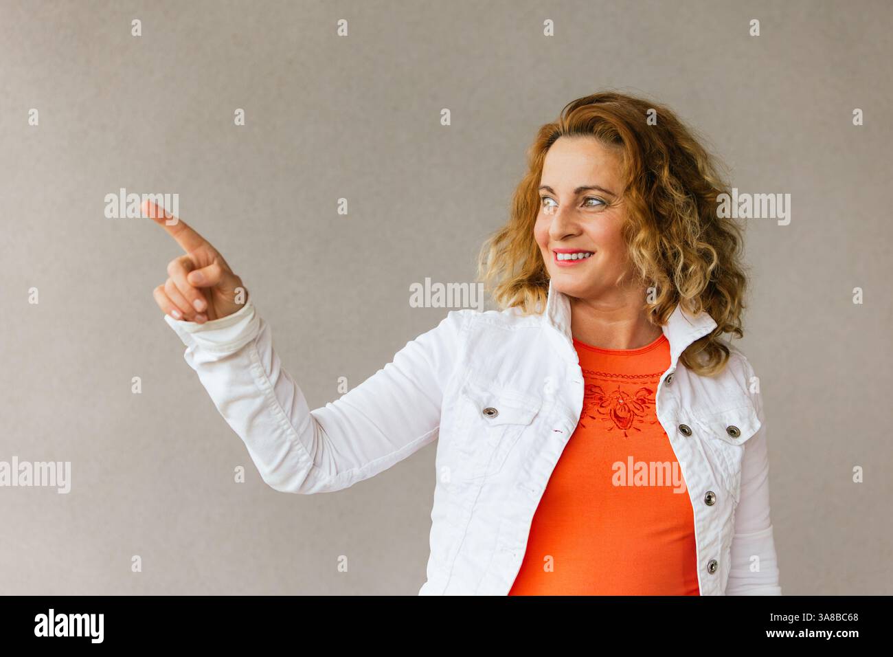 Donna di mezza età con top arancione e giacca bianca rivolta verso il lato con un sorriso allegro, in piedi su uno sfondo neutro in una sicura a Foto Stock
