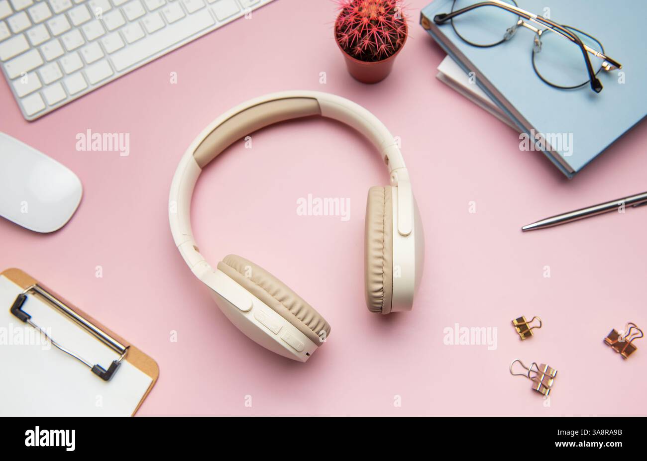 Spazio di lavoro moderno con cuffie wireless, tastiera, mouse, cancelleria, occhiali, e un cactus su sfondo rosa Foto Stock
