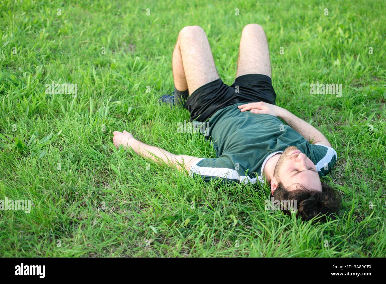 Immagine di un giovane uomo con t-shirt verde sdraiato sull'erba nel parco dopo essersi allenato esausto Foto Stock