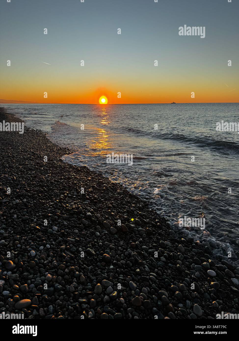 Un tramonto dorato su una spiaggia di ciottoli, con onde delicate che lavano a riva e riflessi scintillanti sull'acqua. Foto Stock