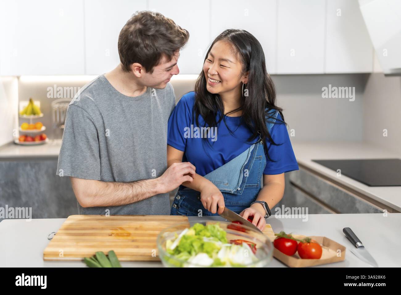 Coppie multietniche che condividono un momento di gioia mentre preparano un pasto in una cucina elegante e moderna. Verdure fresche e un tagliere sono in vista Foto Stock