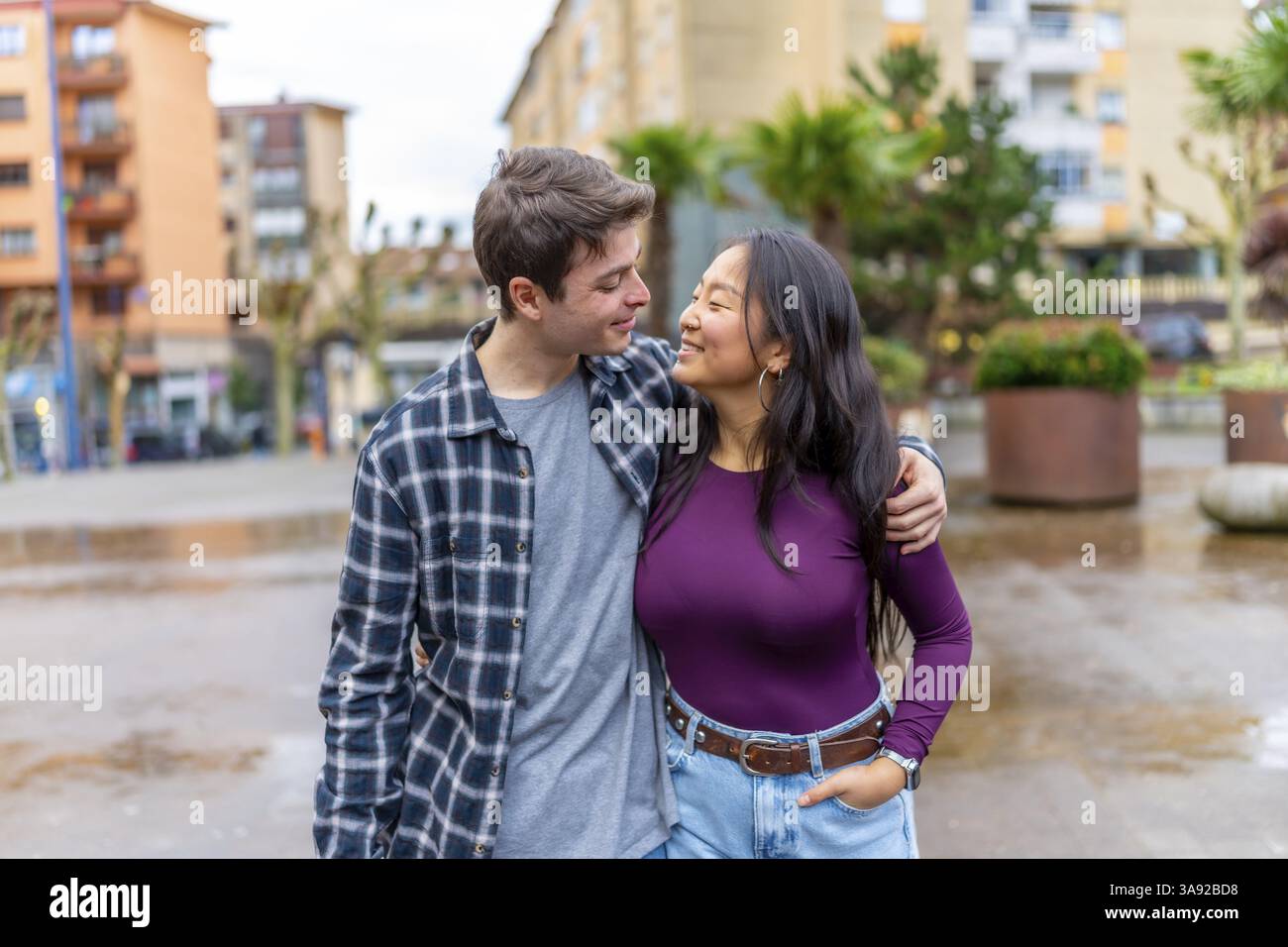 Una felice coppia multietnica condivide un momento di gioia all'aperto, abbracciandosi e sorridendo l'un l'altro, circondati da un ambiente urbano con vegetazione Foto Stock