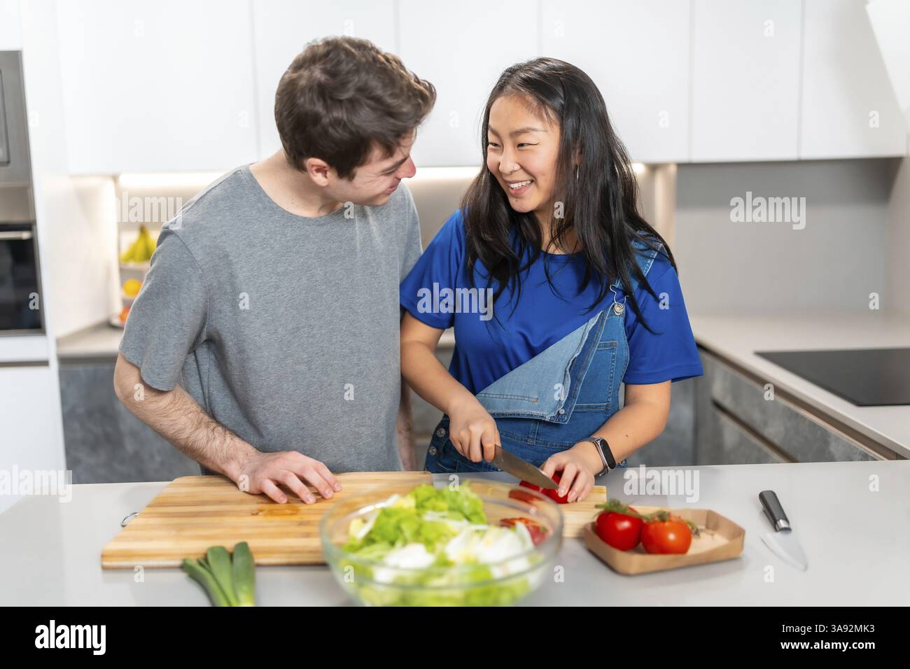 Donna asiatica e uomo caucasico preparano felicemente un pasto in una cucina moderna, mostrando un momento gioioso della vita quotidiana Foto Stock
