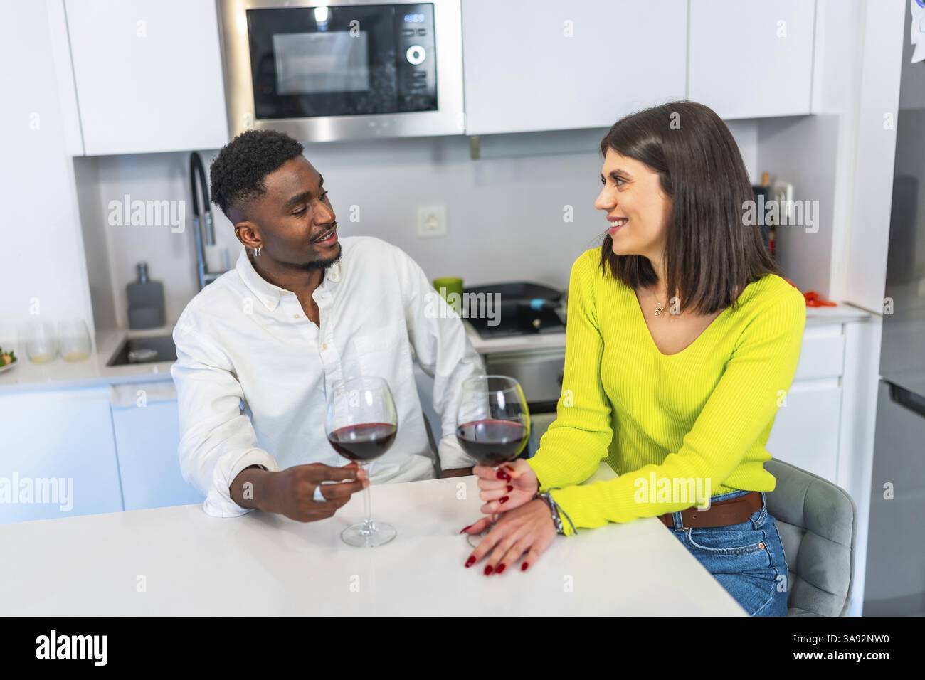 Coppia multietnica che si gode un momento di relax, bevendo vino e conversando mentre prepara un pasto in una cucina contemporanea Foto Stock