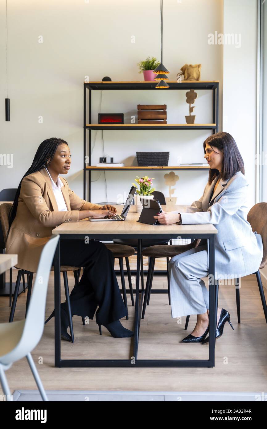 Due donne d'affari stanno lavorando insieme sui loro notebook e tablet nella hall di un hotel, discutendo delle strategie aziendali Foto Stock