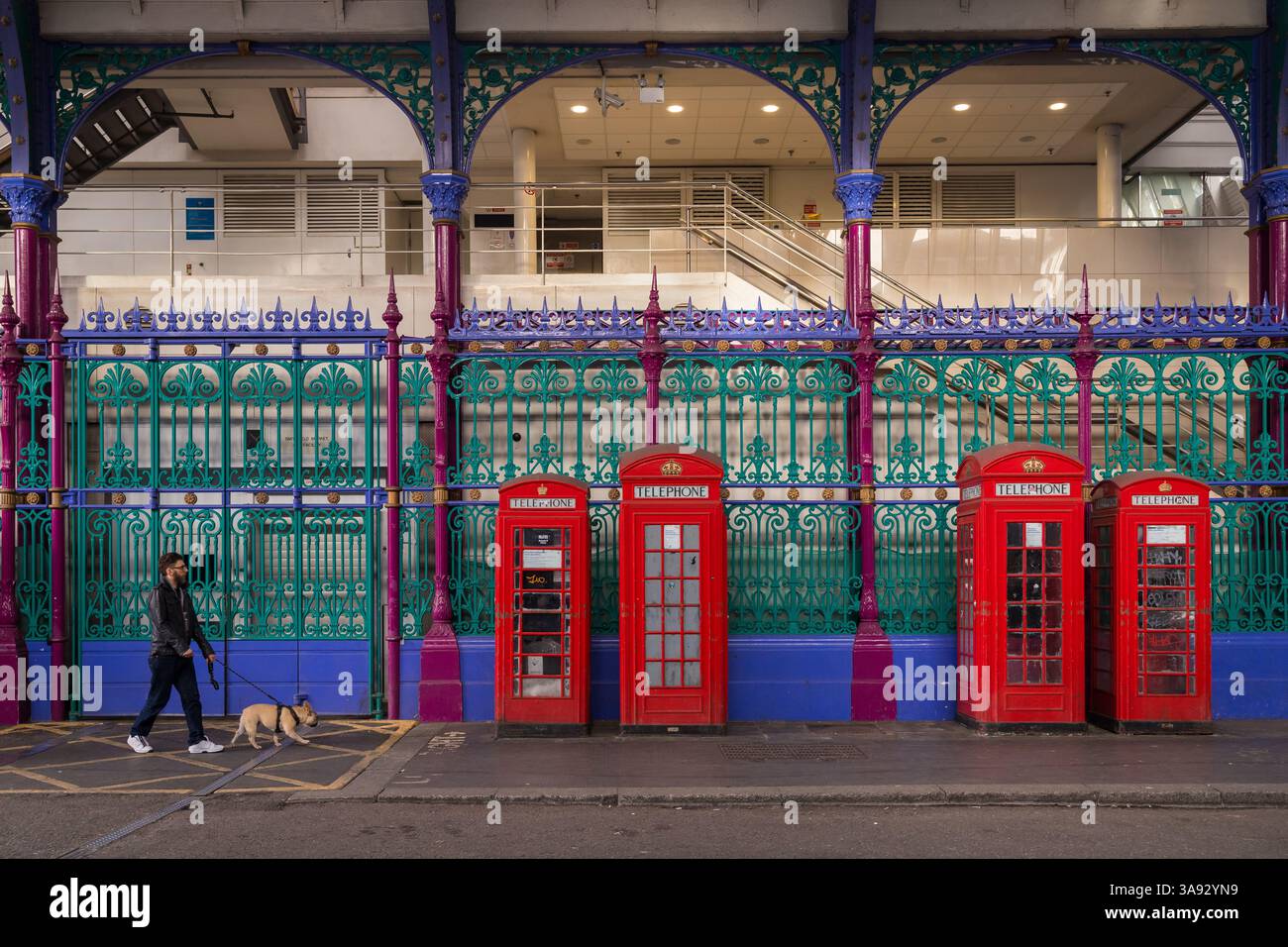 Uomo che cammina con il suo cane vicino alle cabine telefoniche rosse accanto al colorato recinto di Smithfield Market Foto Stock