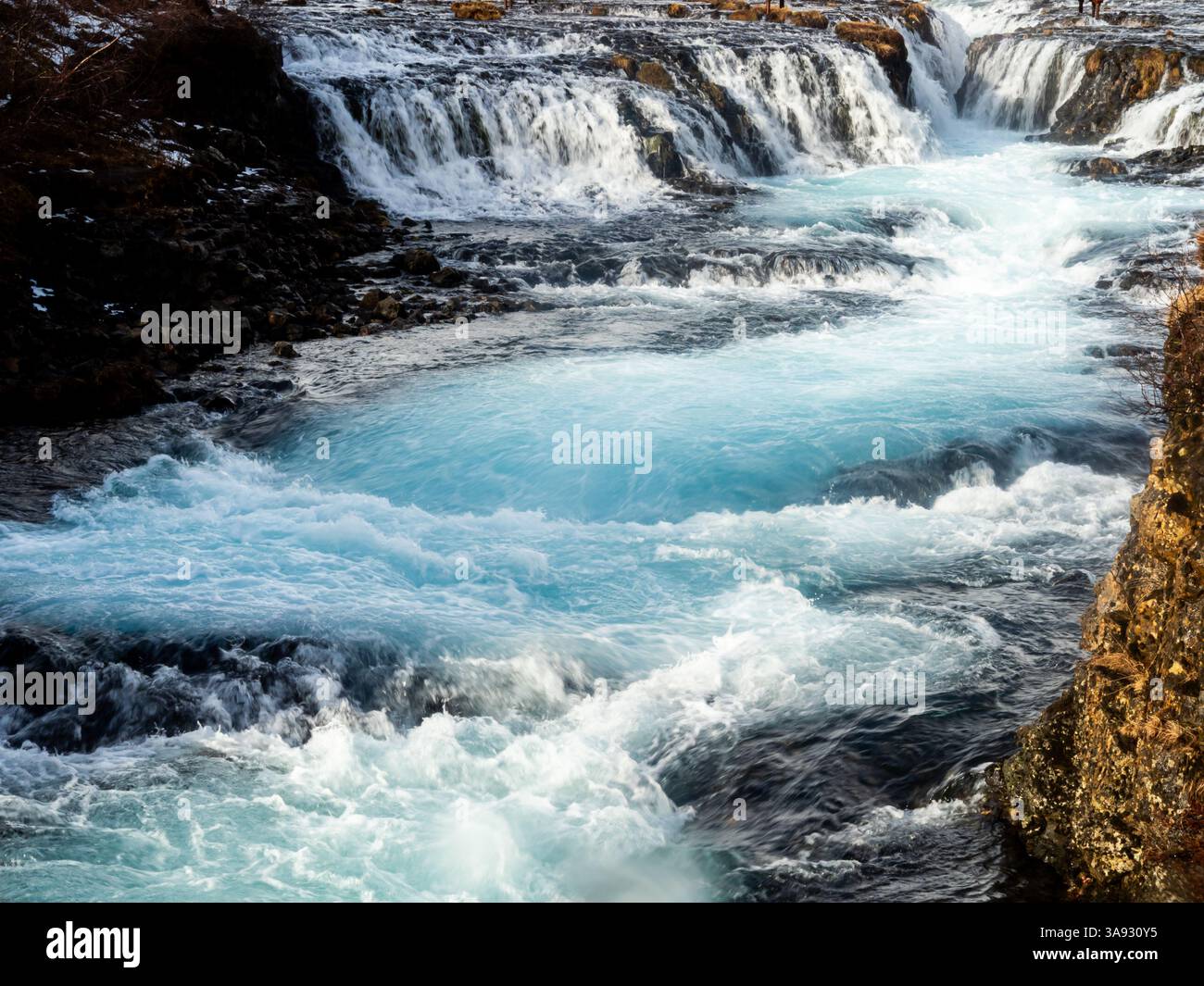 Cascata Bruarfoss, Islanda: Cascate di acque turchesi cristalline sopra l'aspra roccia vulcanica, che creano un contrasto sorprendente tra blu ghiaccio e scuro Foto Stock